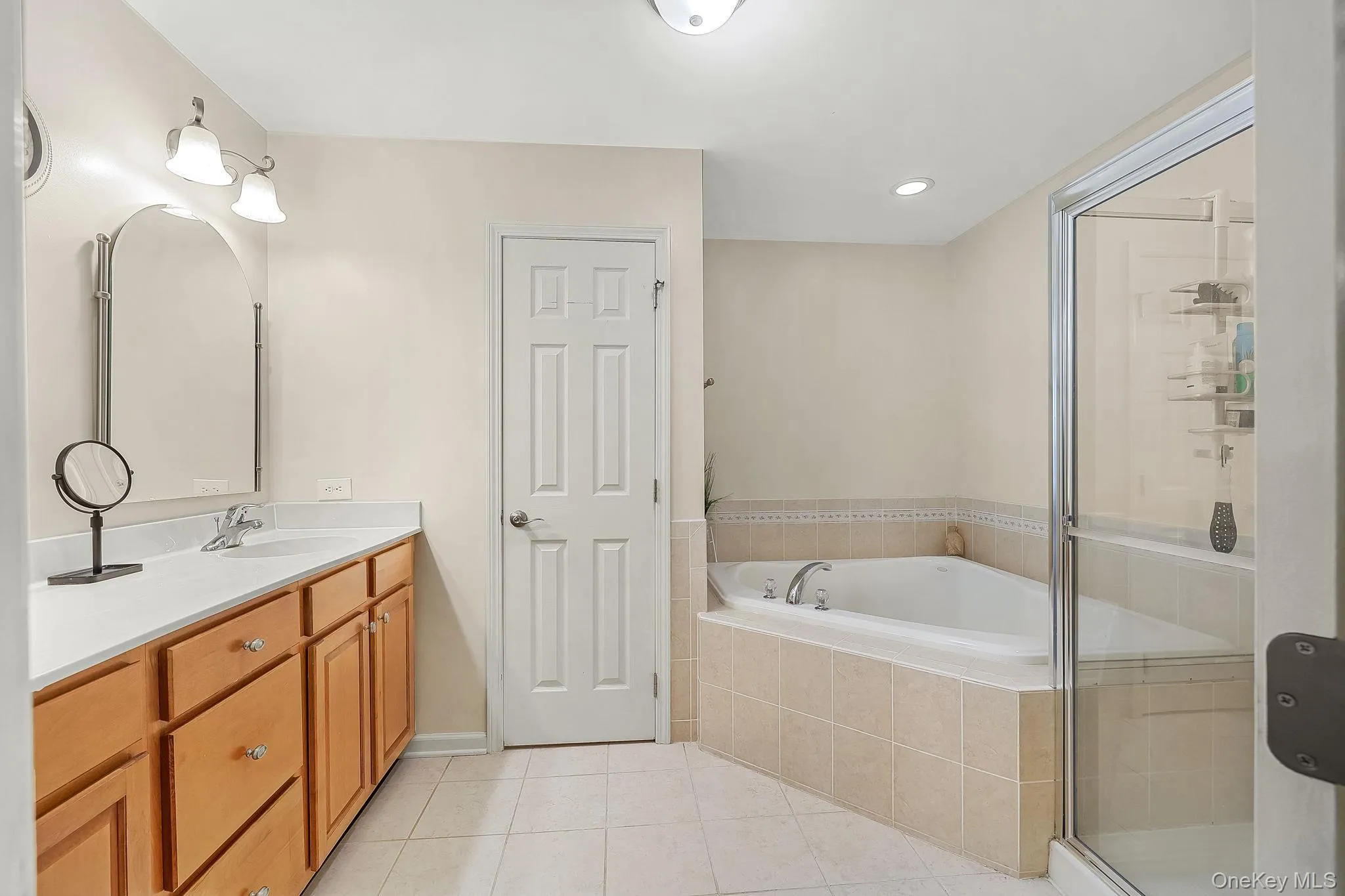 Double vanity with white countertop, soaking tub with tiled surround, and glass-enclosed shower Double vanity with white countertop, soaking tub with tiled surround, and glass-enclosed shower