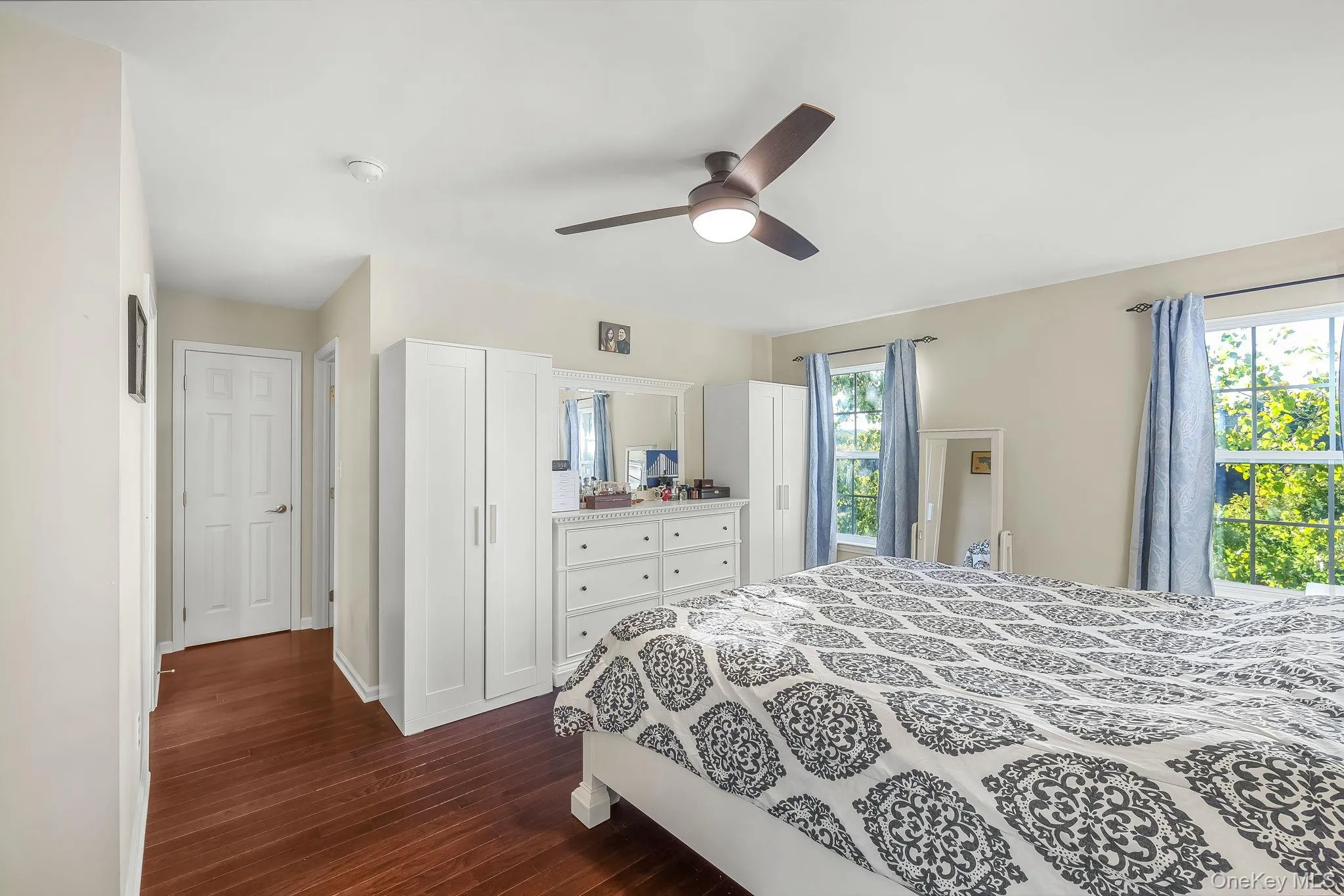 Wood-finish flooring, a ceiling fan with integrated lighting, two windows, and a white paneled door Wood-finish flooring, a ceiling fan with integrated lighting, two windows, and a white paneled door