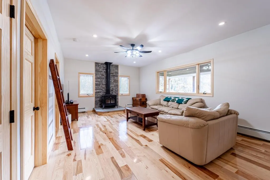Living room featuring light wood-type flooring, baseboard heating, a wood stove, and ceiling fan Living room featuring light wood-type flooring, baseboard heating, a wood stove, and ceiling fan