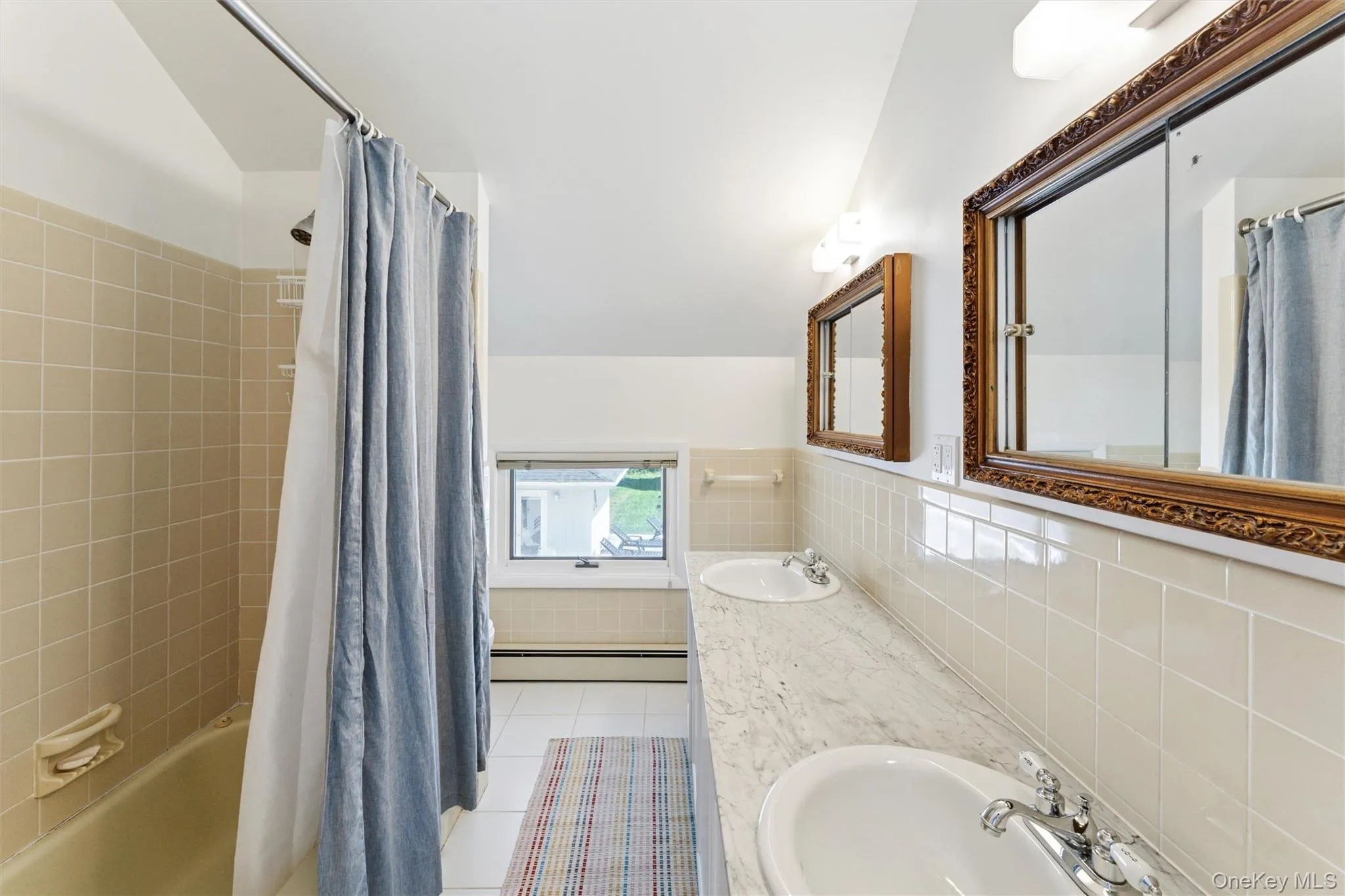 Bathroom featuring light tile patterned floors, shower / bath combination with curtain, tile walls, a baseboard radiator, and double vanity Bathroom featuring light tile patterned floors, shower / bath combination with curtain, tile walls, a baseboard radiator, and double vanity