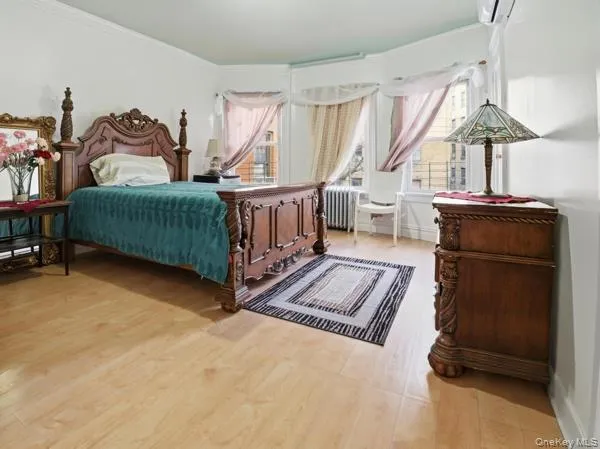 Bedroom featuring multiple windows, ornamental molding, a wall unit AC, light wood-type flooring, and radiator heating unit Bedroom featuring multiple windows, ornamental molding, a wall unit AC, light wood-type flooring, and radiator heating unit