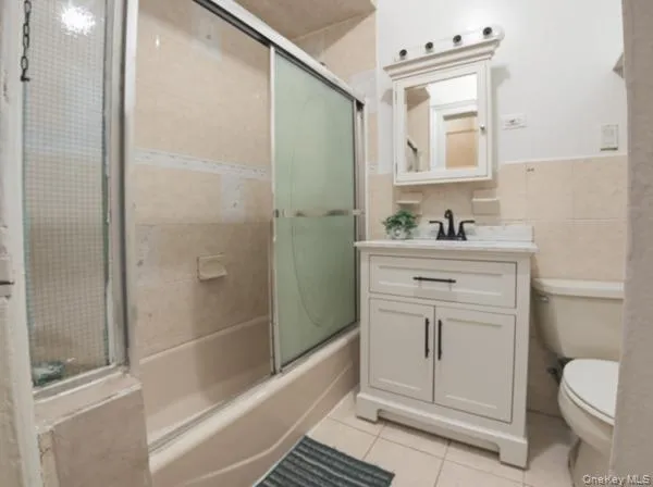 Full bathroom featuring vanity, bath / shower combo with glass door, light tile patterned floors, tile walls, and wainscoting Full bathroom featuring vanity, bath / shower combo with glass door, light tile patterned floors, tile walls, and wainscoting
