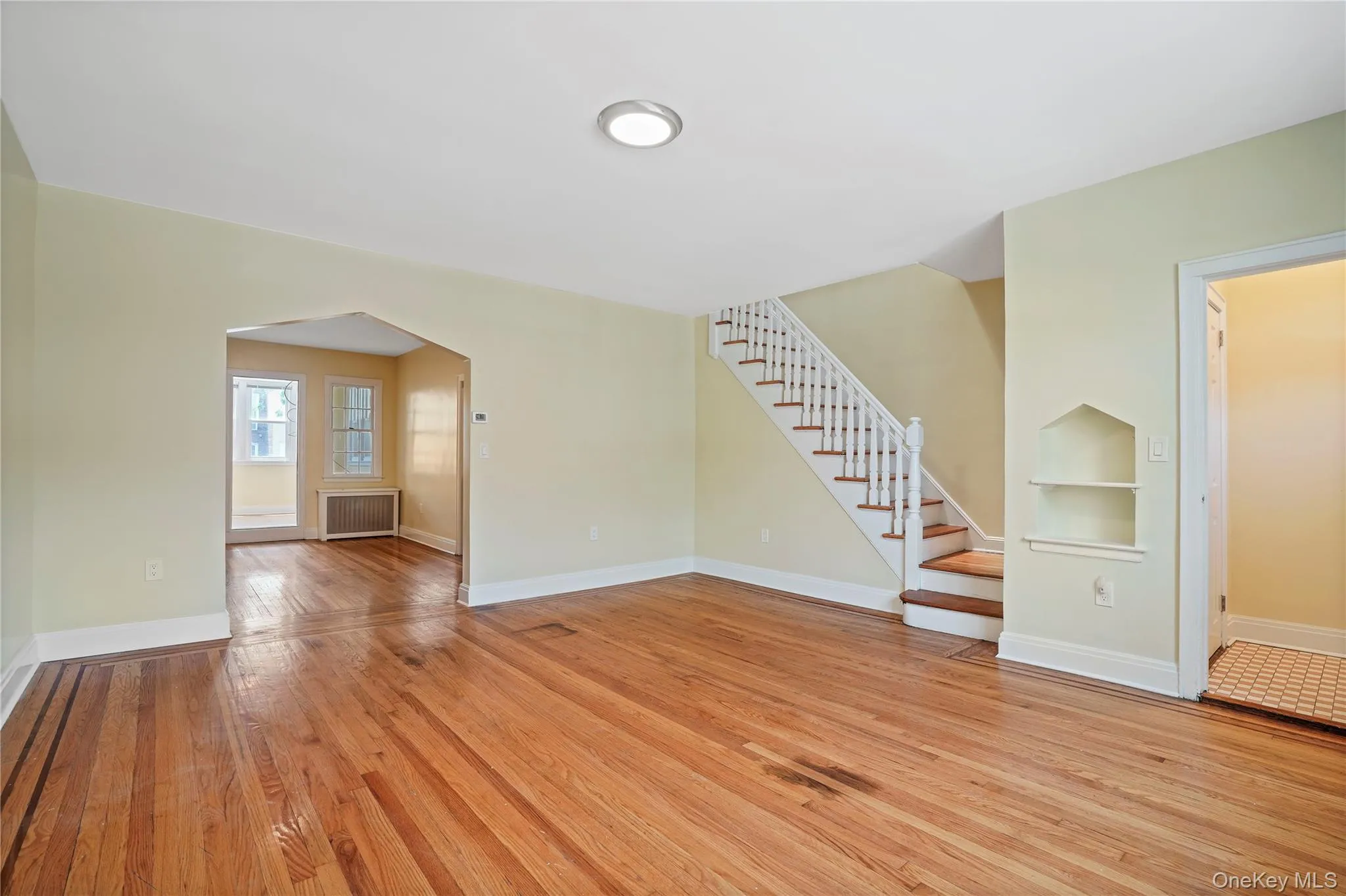 Unfurnished living room with arched walkways, radiator, light wood finished floors, stairs, and built in features Unfurnished living room with arched walkways, radiator, light wood finished floors, stairs, and built in features