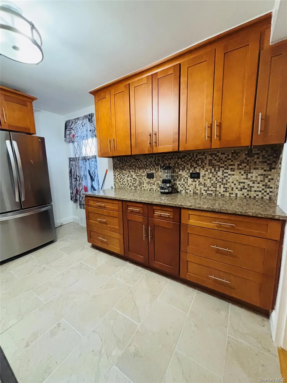 Kitchen with tasteful backsplash, brown cabinetry, freestanding refrigerator, light stone countertops, and baseboards Kitchen with tasteful backsplash, brown cabinetry, freestanding refrigerator, light stone countertops, and baseboards