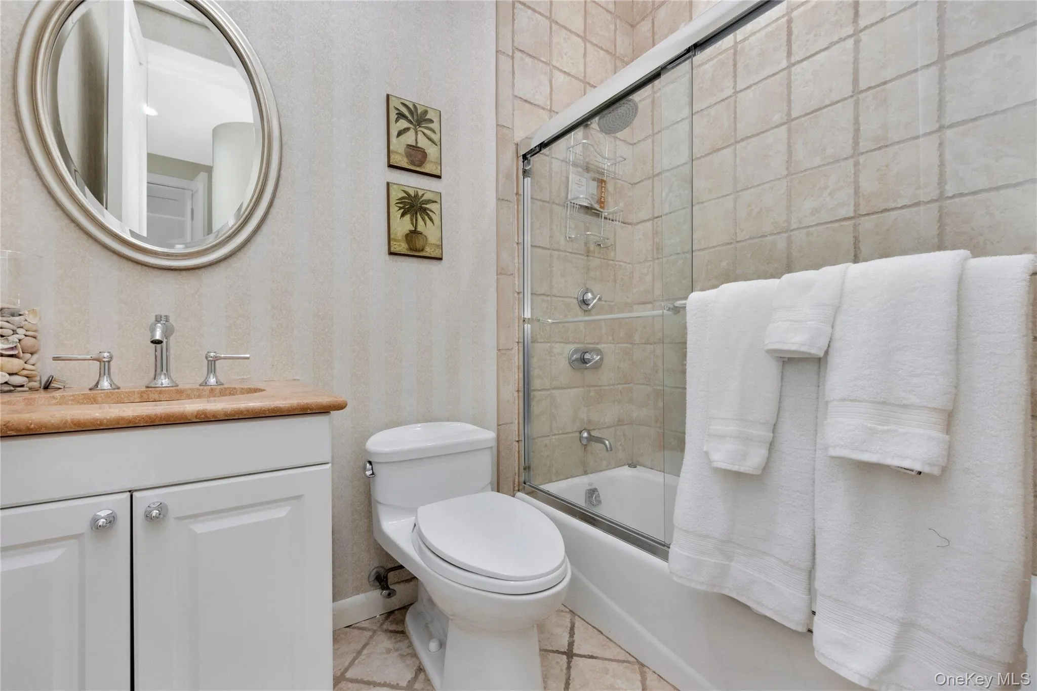 Bathroom with shower / bath combination with glass door, toilet, vanity, tile patterned flooring, and wallpapered walls Bathroom with shower / bath combination with glass door, toilet, vanity, tile patterned flooring, and wallpapered walls