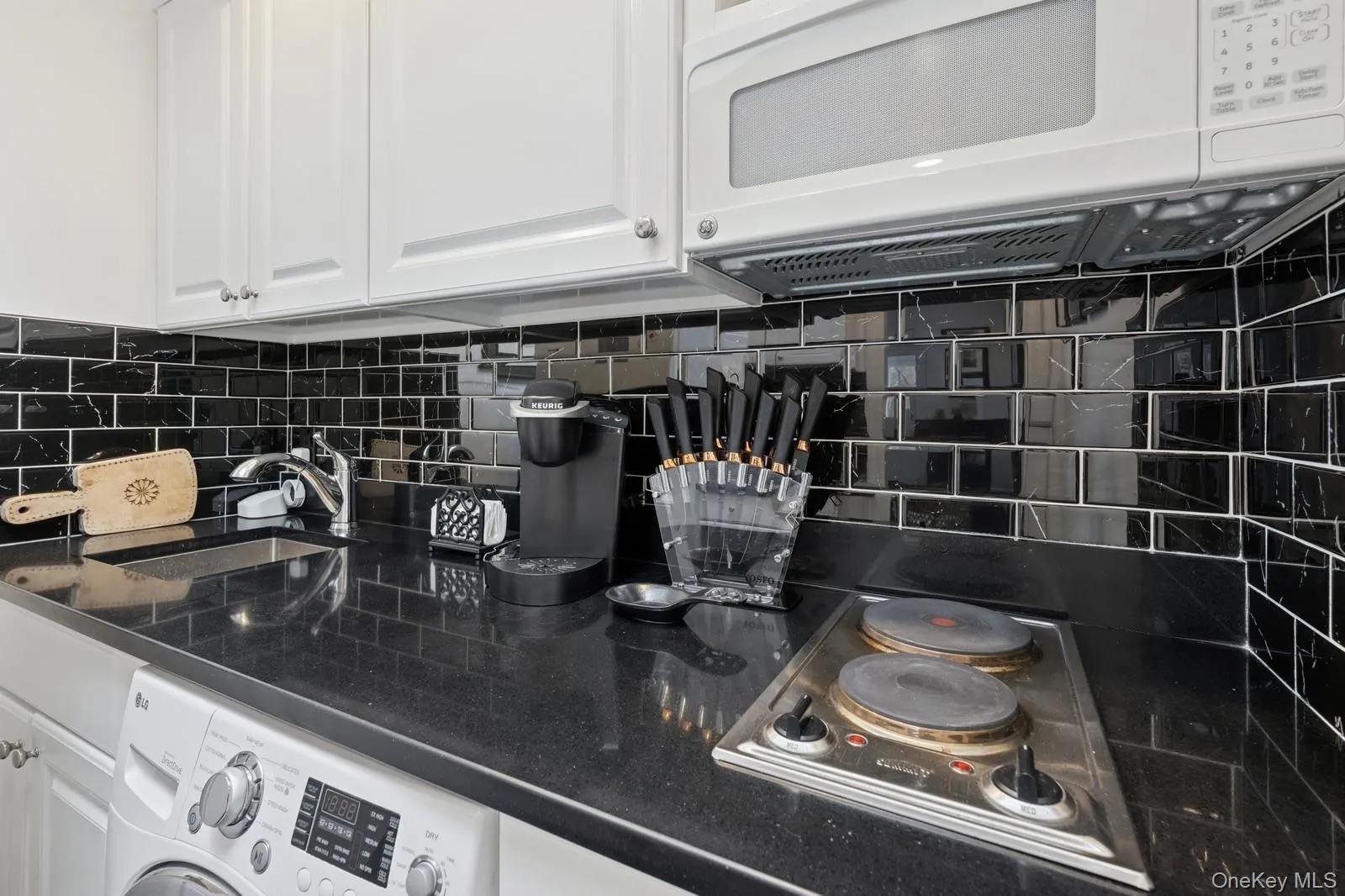 Kitchen featuring washer / clothes dryer, electric cooktop, decorative backsplash, white microwave, and white cabinets Kitchen featuring washer / clothes dryer, electric cooktop, decorative backsplash, white microwave, and white cabinets