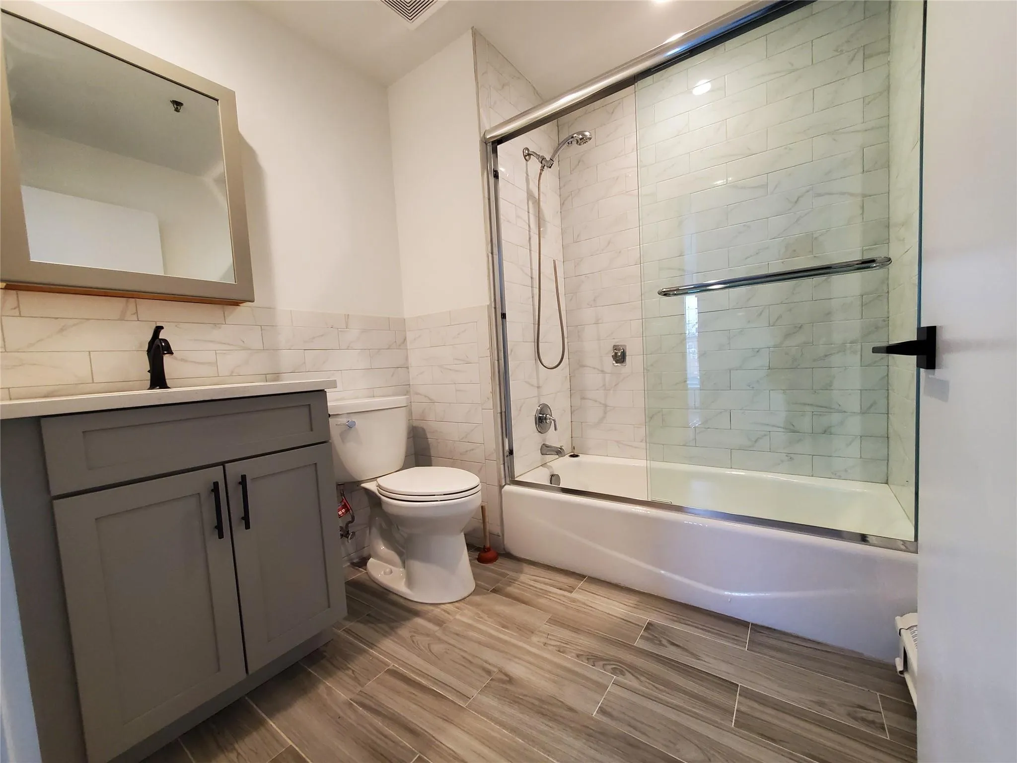 Bathroom featuring toilet, vanity, bath / shower combo with glass door and tile walls Bathroom featuring toilet, vanity, bath / shower combo with glass door and tile walls
