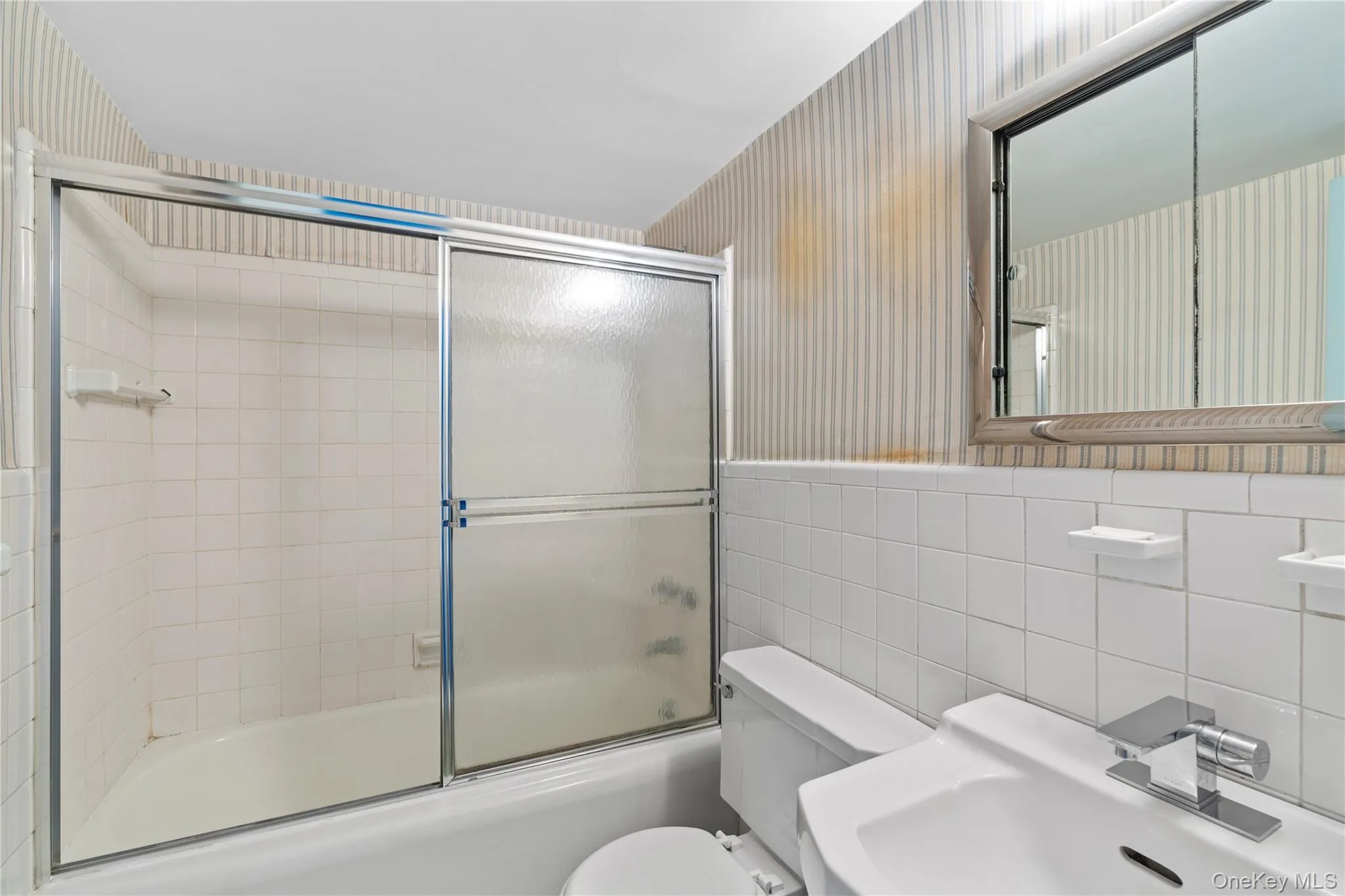 Full bath with toilet, a sink, tile walls, wallpapered walls, and bath / shower combo with glass door Full bath with toilet, a sink, tile walls, wallpapered walls, and bath / shower combo with glass door