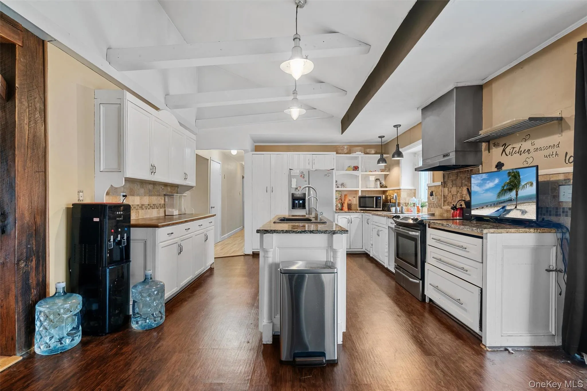Kitchen featuring a center island with sink, open shelves, lofted ceiling with beams, stainless steel appliances, and wall chimney range hood Kitchen featuring a center island with sink, open shelves, lofted ceiling with beams, stainless steel appliances, and wall chimney range hood