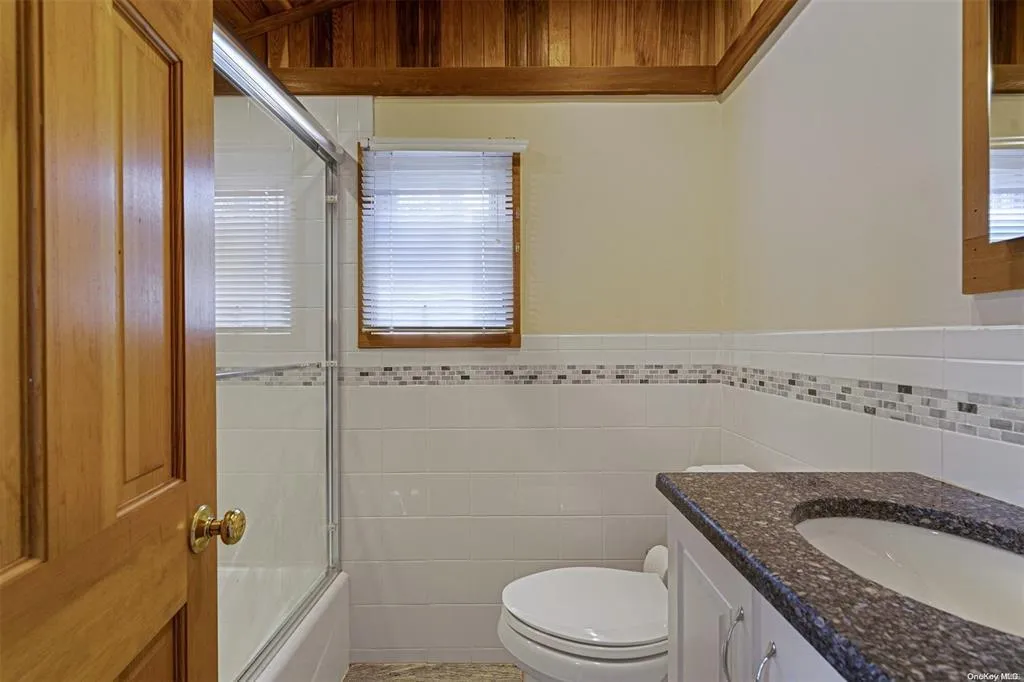 Bathroom featuring wainscoting, toilet, combined bath / shower with glass door, vanity, and tile walls Bathroom featuring wainscoting, toilet, combined bath / shower with glass door, vanity, and tile walls