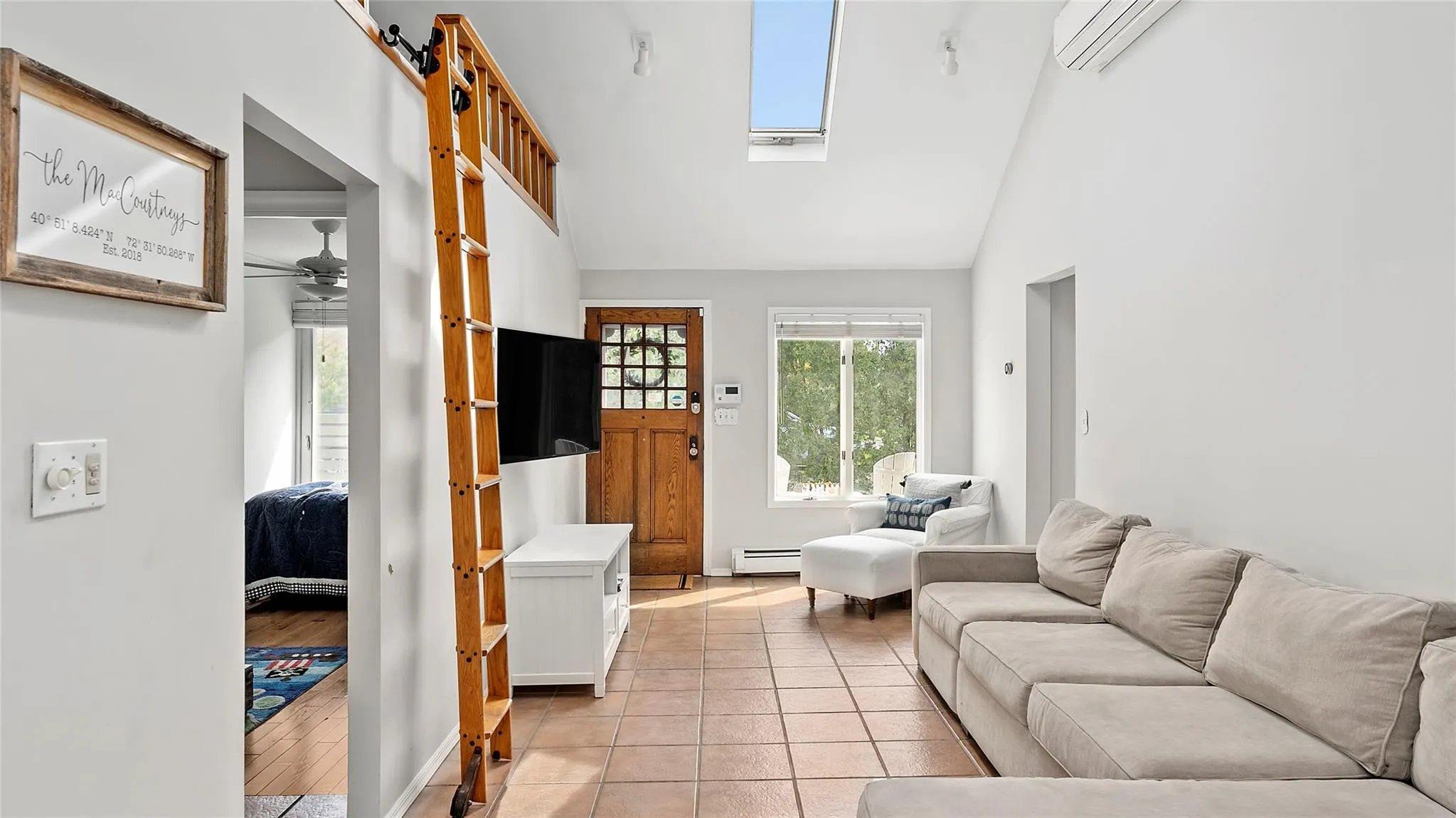 Living room featuring light tile patterned floors, a ceiling fan, an AC wall unit, vaulted ceiling with skylight, and baseboards Living room featuring light tile patterned floors, a ceiling fan, an AC wall unit, vaulted ceiling with skylight, and baseboards