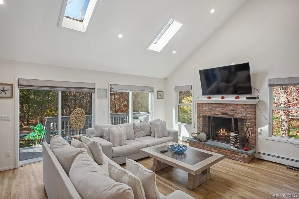 Living room featuring high vaulted ceiling, a skylight, light wood-style floors, a brick fireplace, and baseboard heating Living room featuring high vaulted ceiling, a skylight, light wood-style floors, a brick fireplace, and baseboard heating