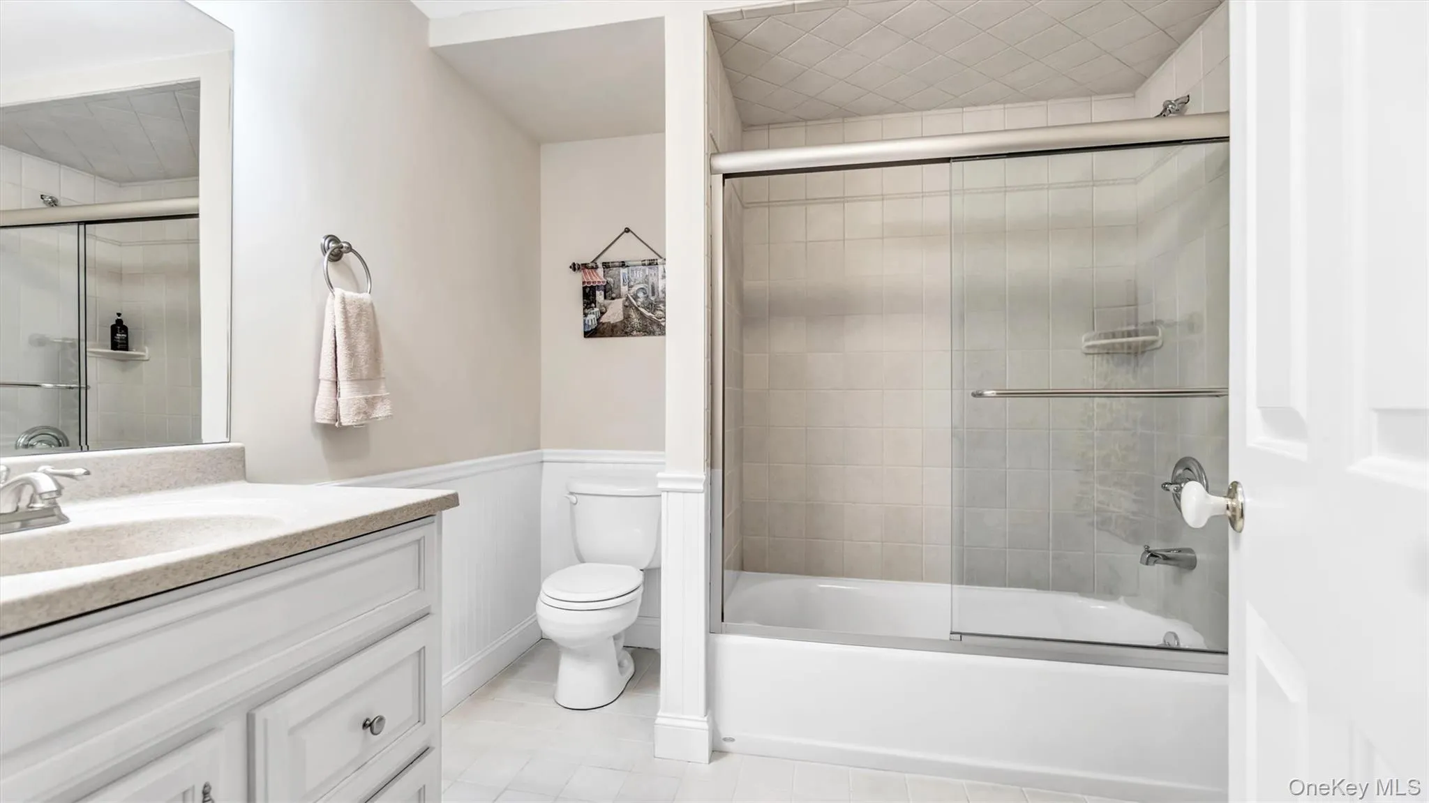 Bathroom with tile patterned floors, a wainscoted wall, toilet, bath / shower combo with glass door, and vanity Bathroom with tile patterned floors, a wainscoted wall, toilet, bath / shower combo with glass door, and vanity