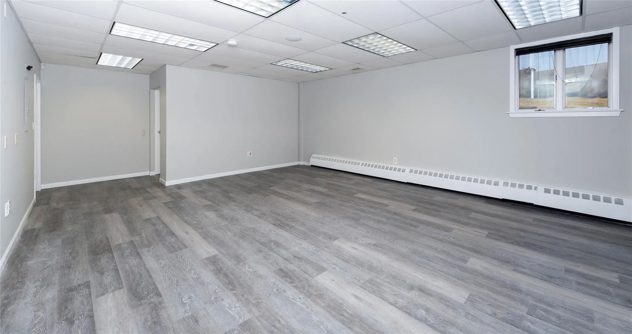 Unfurnished room featuring baseboard heating, a drop ceiling, baseboards, and wood finished floors Unfurnished room featuring baseboard heating, a drop ceiling, baseboards, and wood finished floors