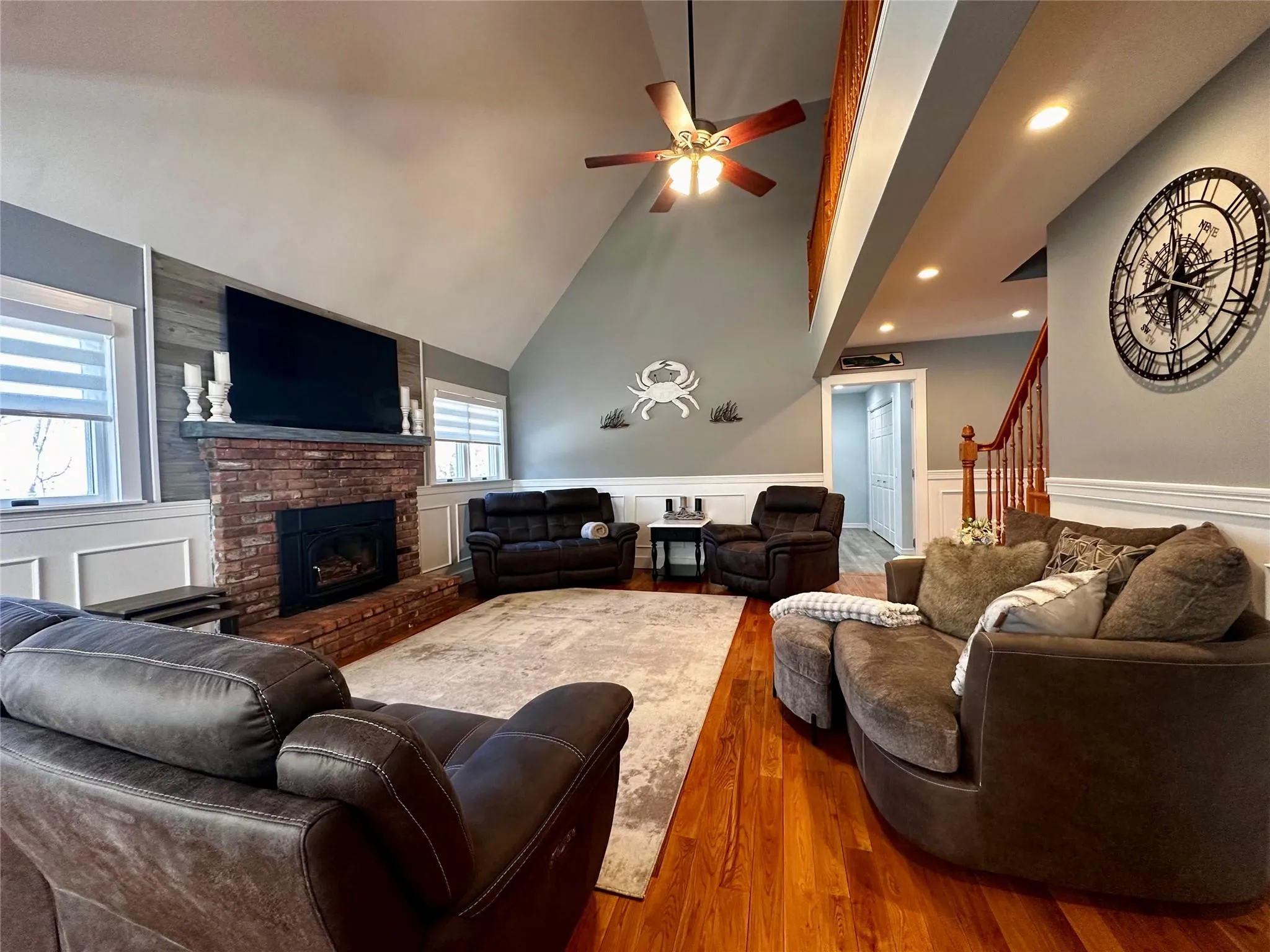 Living room featuring a wainscoted wall, a brick fireplace, stairs, wood finished floors, and a ceiling fan Living room featuring a wainscoted wall, a brick fireplace, stairs, wood finished floors, and a ceiling fan