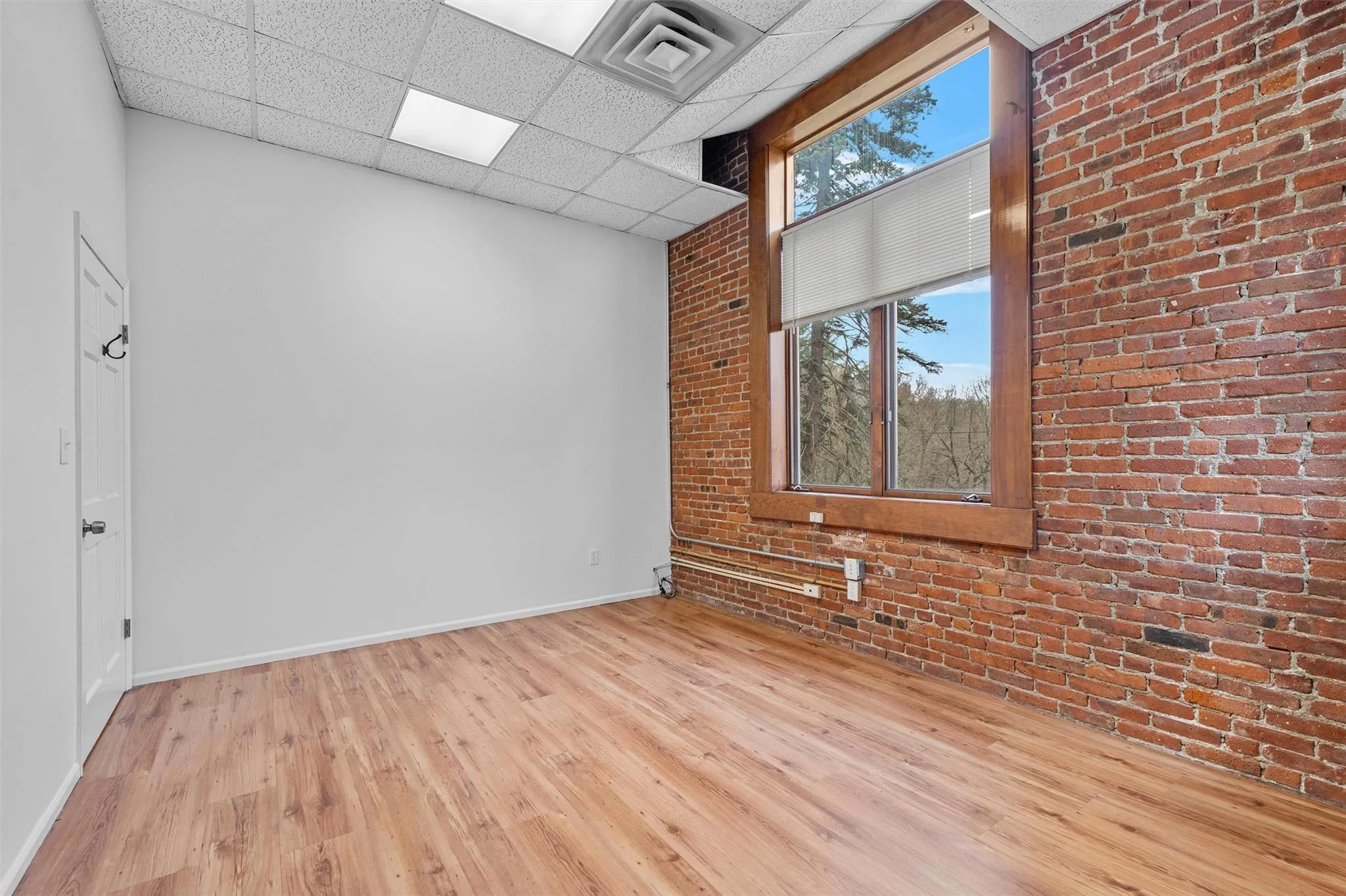 Unfurnished room featuring visible vents, baseboards, wood finished floors, brick wall, and a paneled ceiling Unfurnished room featuring visible vents, baseboards, wood finished floors, brick wall, and a paneled ceiling