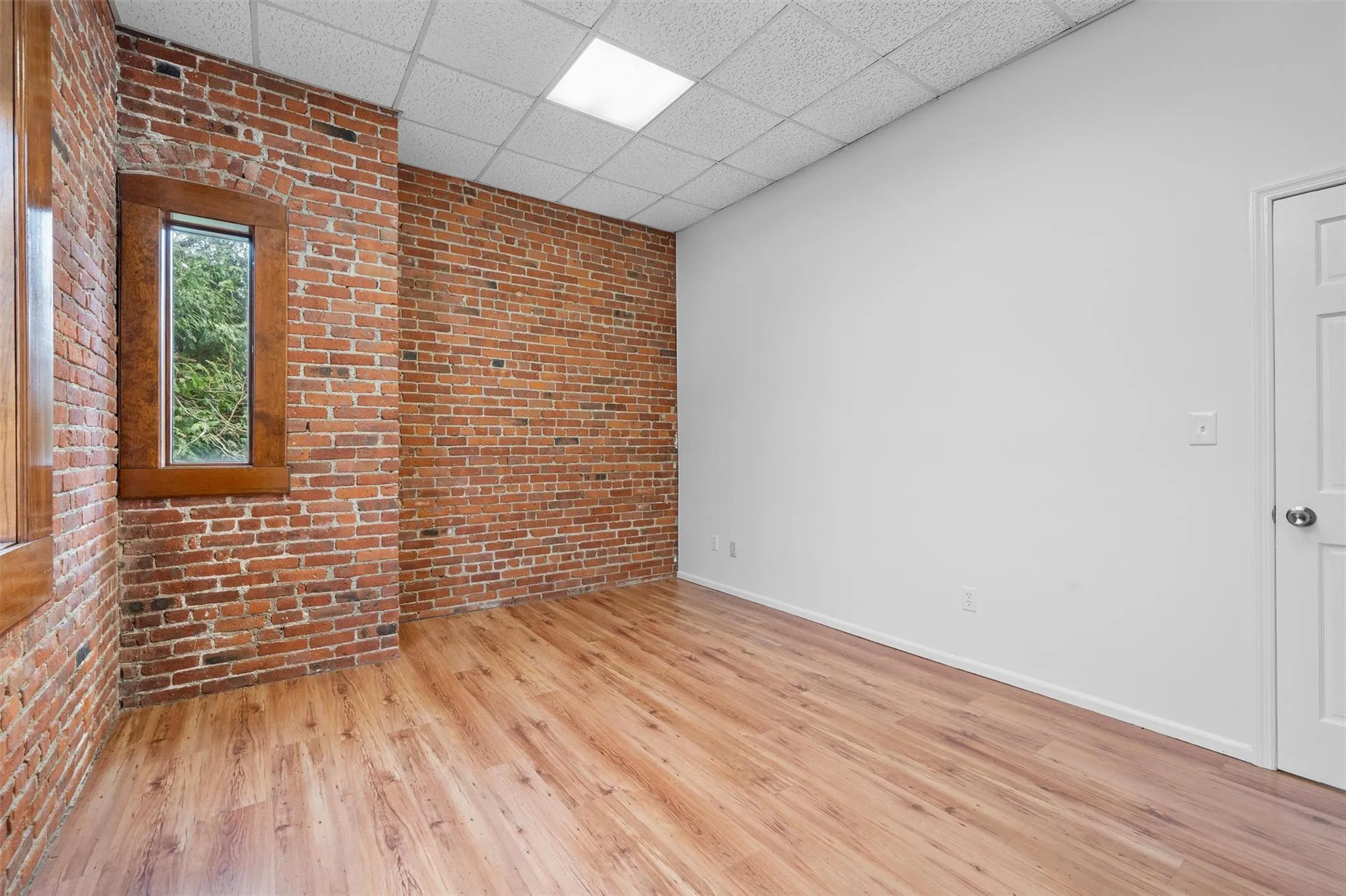 Empty room featuring baseboards, wood finished floors, a paneled ceiling, and brick wall Empty room featuring baseboards, wood finished floors, a paneled ceiling, and brick wall