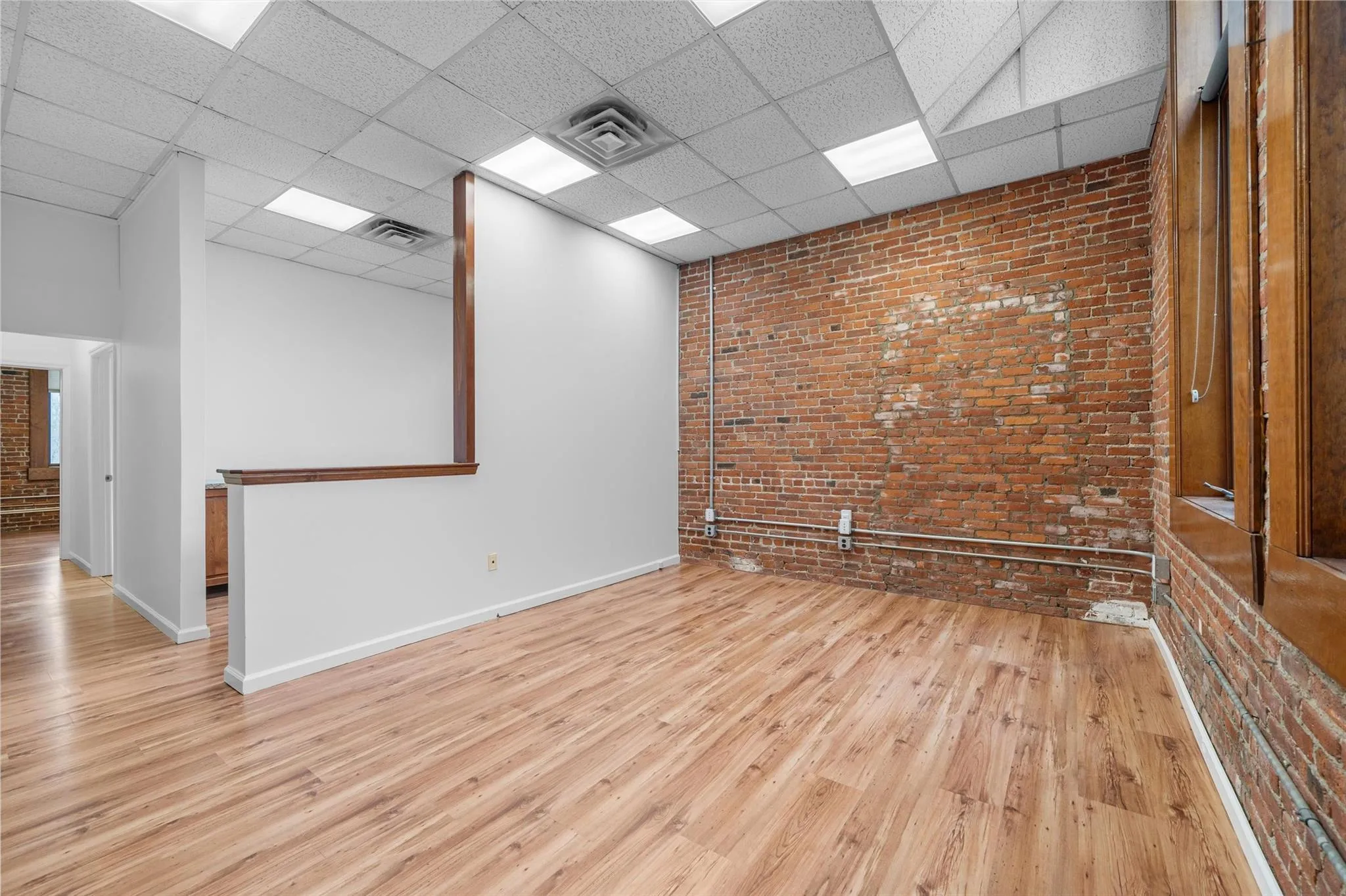 Empty room with a paneled ceiling, baseboards, brick wall, and wood finished floors Empty room with a paneled ceiling, baseboards, brick wall, and wood finished floors