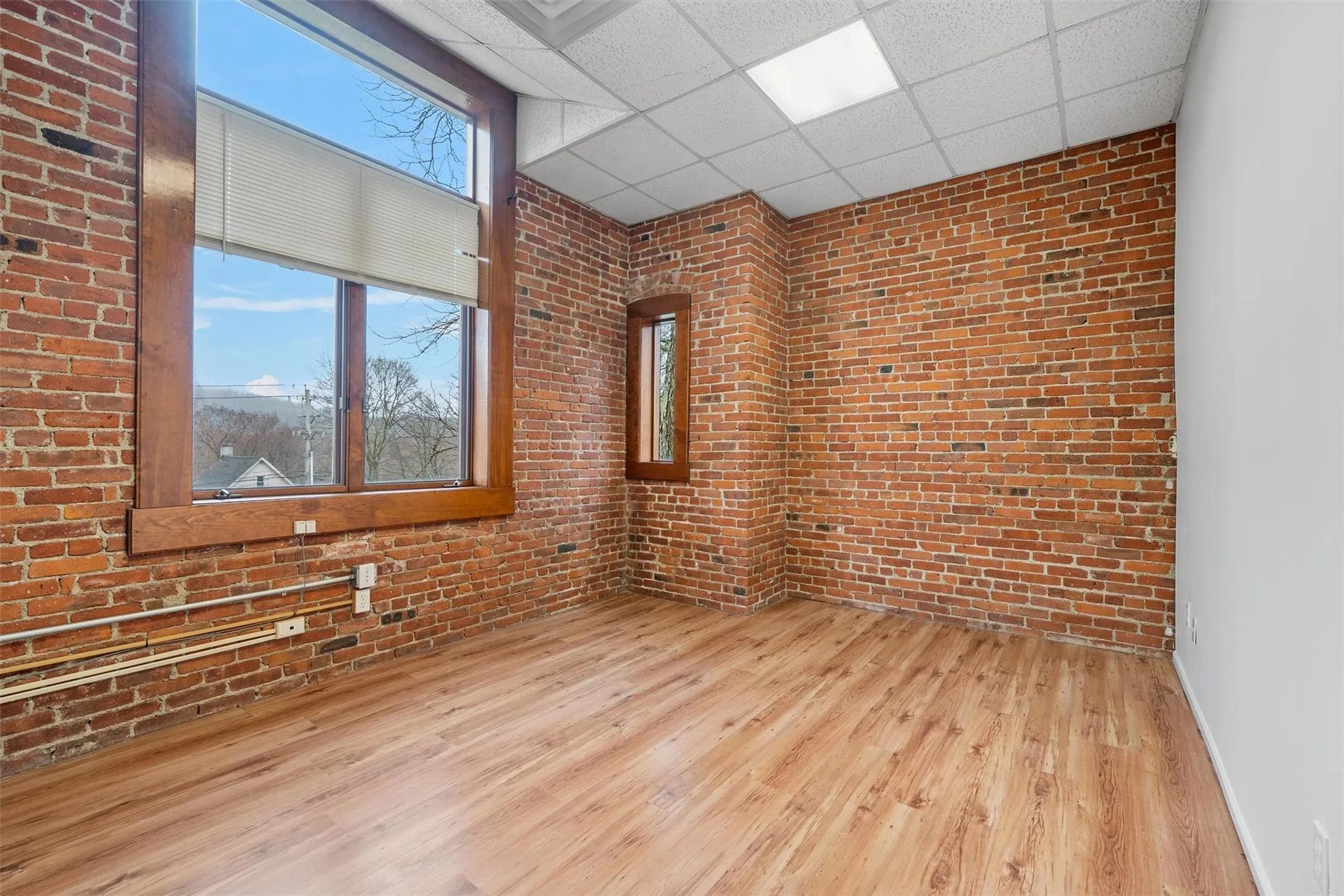 Empty room with baseboards, a drop ceiling, brick wall, and wood finished floors Empty room with baseboards, a drop ceiling, brick wall, and wood finished floors