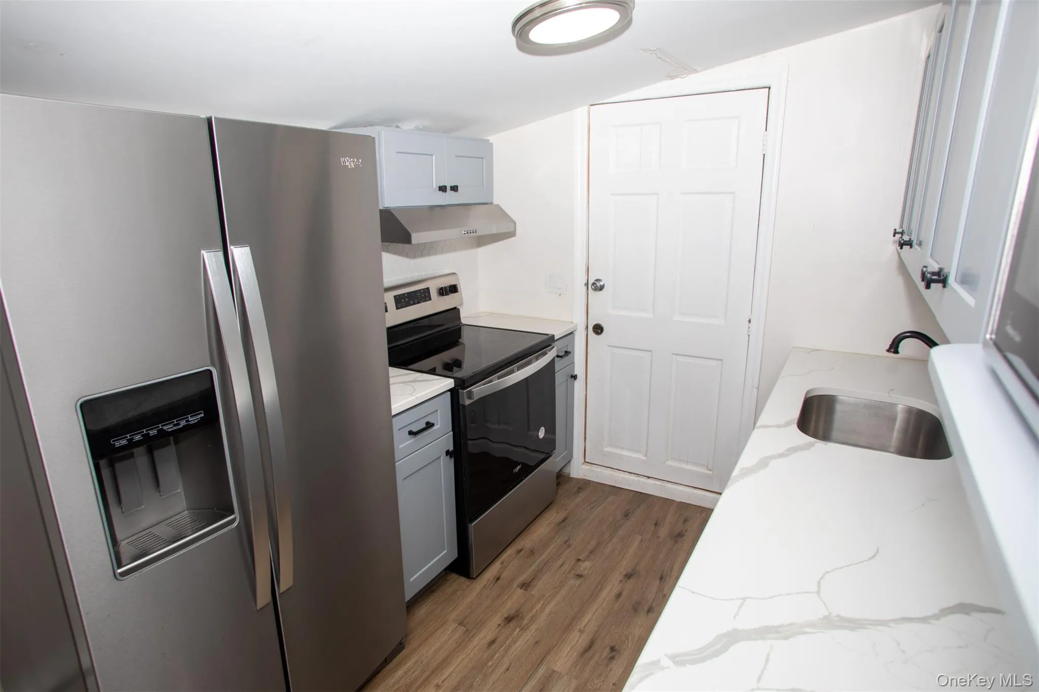Kitchen featuring wood finished floors, appliances with stainless steel finishes, under cabinet range hood, a sink, and light stone counters Kitchen featuring wood finished floors, appliances with stainless steel finishes, under cabinet range hood, a sink, and light stone counters