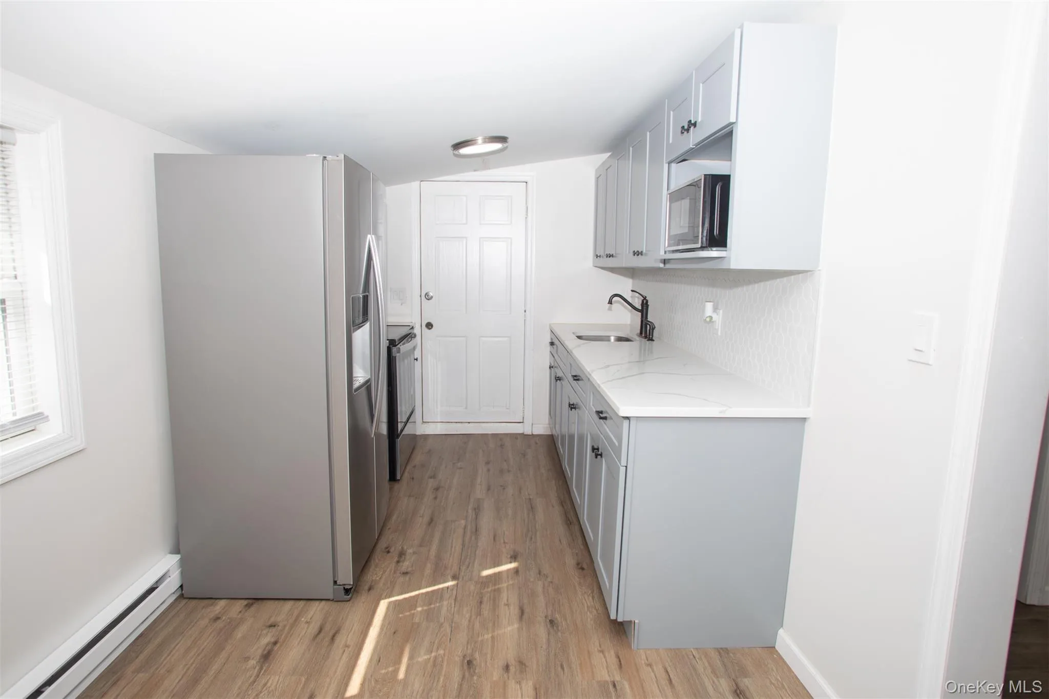 Kitchen featuring stainless steel refrigerator with ice dispenser, a baseboard heating unit, a sink, light wood finished floors, and light stone countertops Kitchen featuring stainless steel refrigerator with ice dispenser, a baseboard heating unit, a sink, light wood finished floors, and light stone countertops
