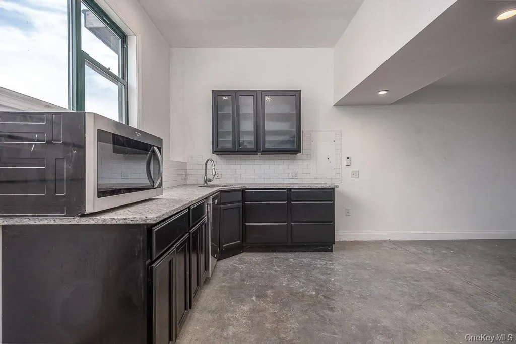 Kitchen featuring stainless steel microwave, concrete flooring, glass insert cabinets, and backsplash Kitchen featuring stainless steel microwave, concrete flooring, glass insert cabinets, and backsplash