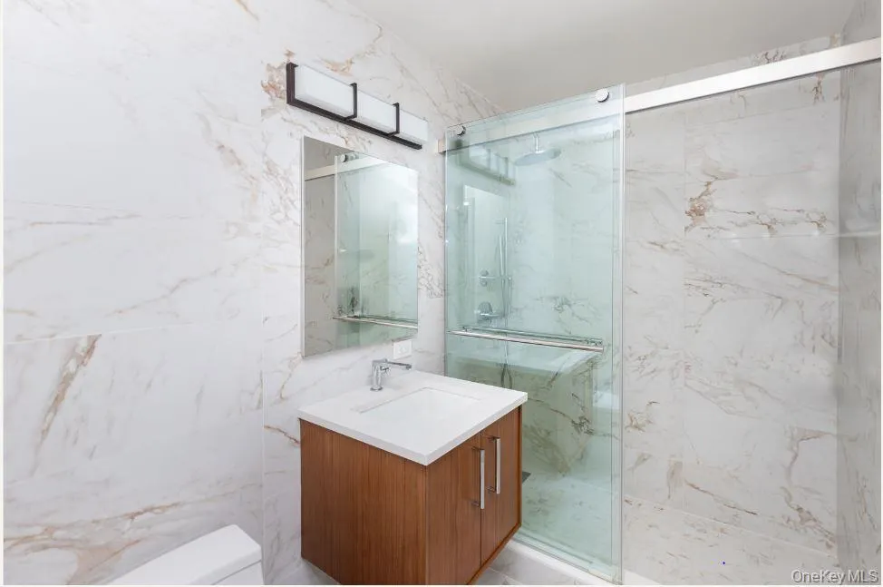 Bathroom featuring a marble finish shower, tile walls, toilet, and vanity Bathroom featuring a marble finish shower, tile walls, toilet, and vanity