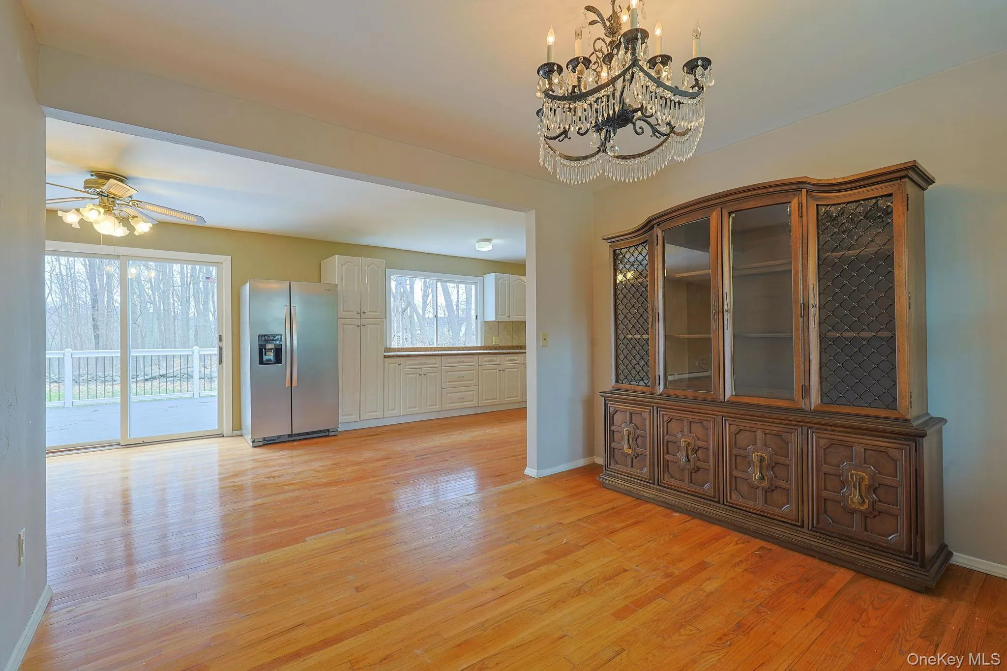 Hardwood floors extend through this room, featuring a chandelier, and a view into a kitchen with light-colored cabinetry and a stainless steel refrigerator Hardwood floors extend through this room, featuring a chandelier, and a view into a kitchen with light-colored cabinetry and a stainless steel refrigerator