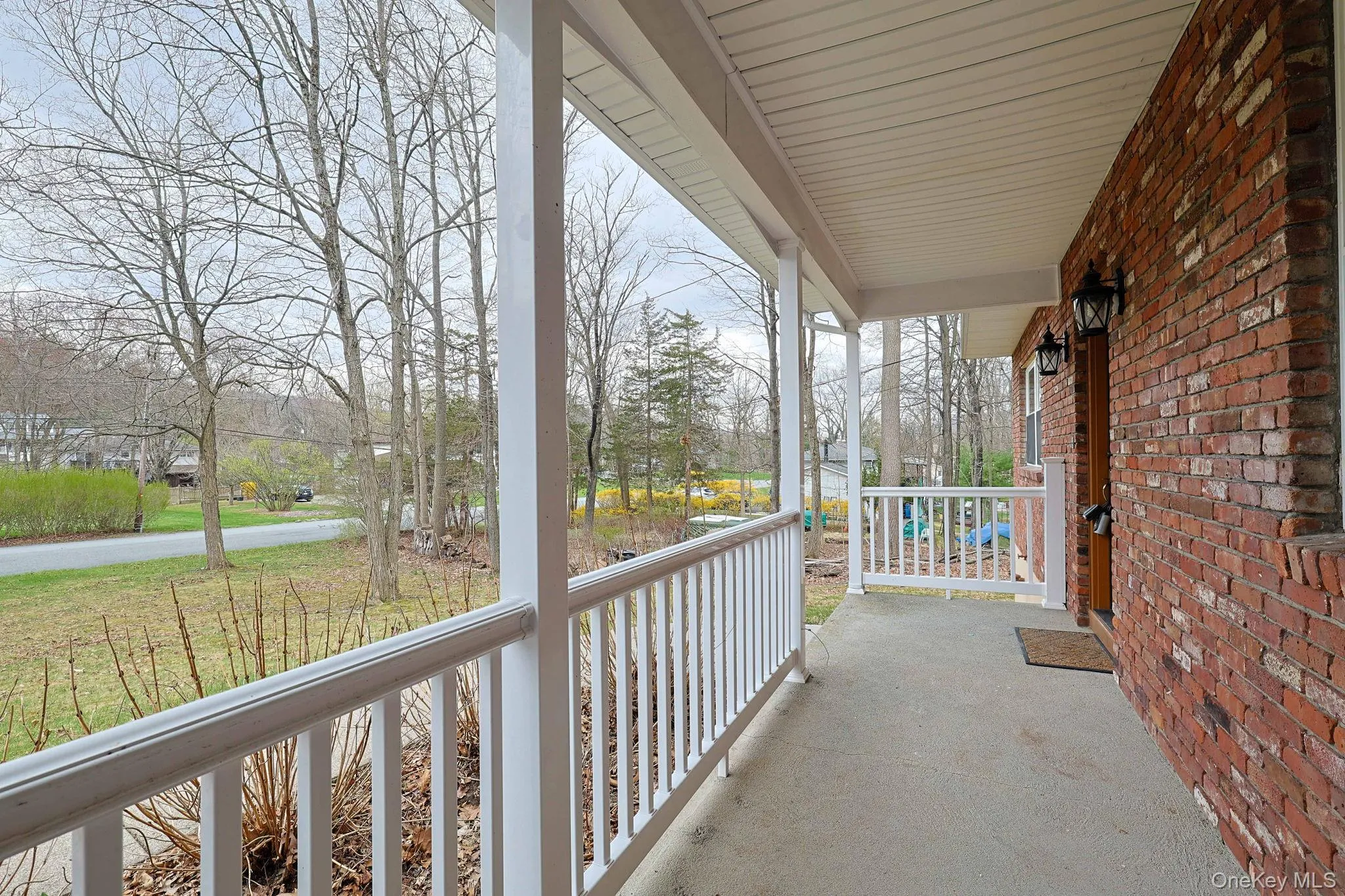 Inviting covered front porch with white railings and brick exterior, offering views of the surrounding trees and street Inviting covered front porch with white railings and brick exterior, offering views of the surrounding trees and street