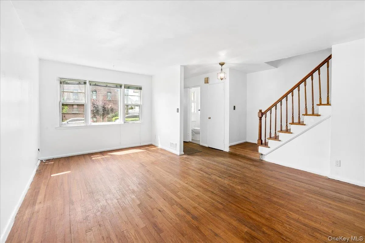 Spacious living area featuring wood-finish flooring, white walls, and a prominent staircase with wood railings Spacious living area featuring wood-finish flooring, white walls, and a prominent staircase with wood railings