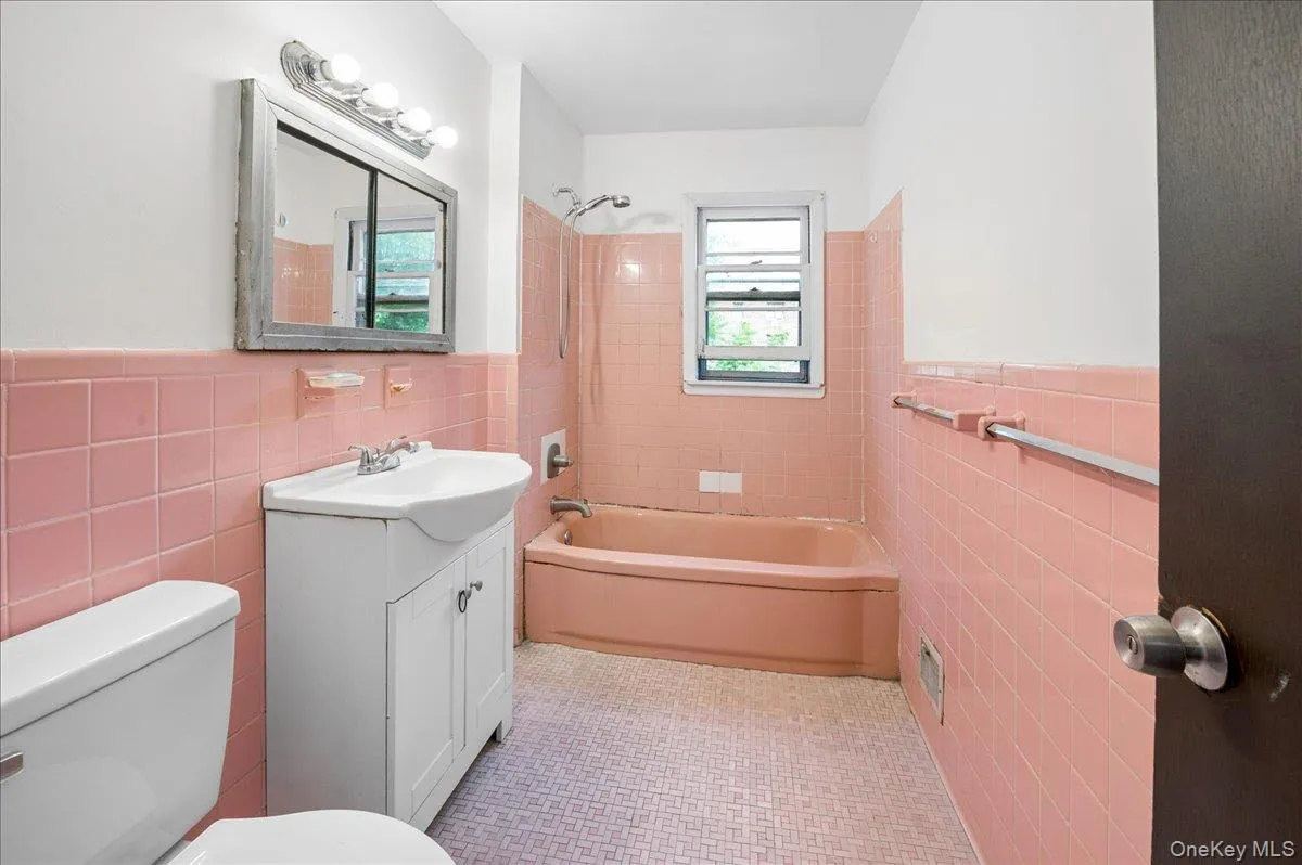 Bathroom featuring pink wall tiling, a matching pink bathtub, a white vanity with integrated sink, a mirrored medicine cabinet, and a window Bathroom featuring pink wall tiling, a matching pink bathtub, a white vanity with integrated sink, a mirrored medicine cabinet, and a window