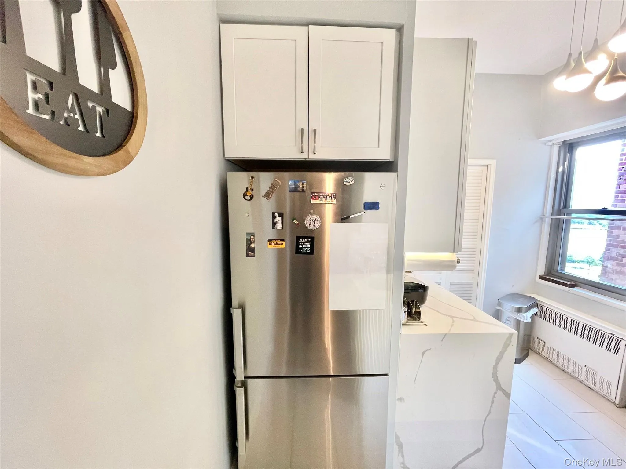 Kitchen featuring radiator, freestanding refrigerator, white cabinetry, light stone countertops, and decorative light fixtures Kitchen featuring radiator, freestanding refrigerator, white cabinetry, light stone countertops, and decorative light fixtures