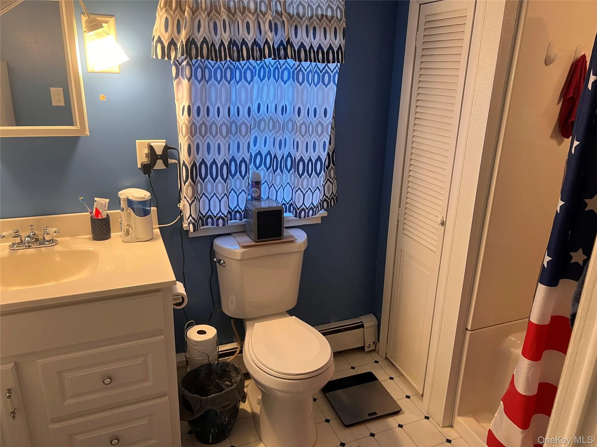 Bathroom with curtained shower, vanity, baseboard heating, light tile patterned floors, and a closet Bathroom with curtained shower, vanity, baseboard heating, light tile patterned floors, and a closet
