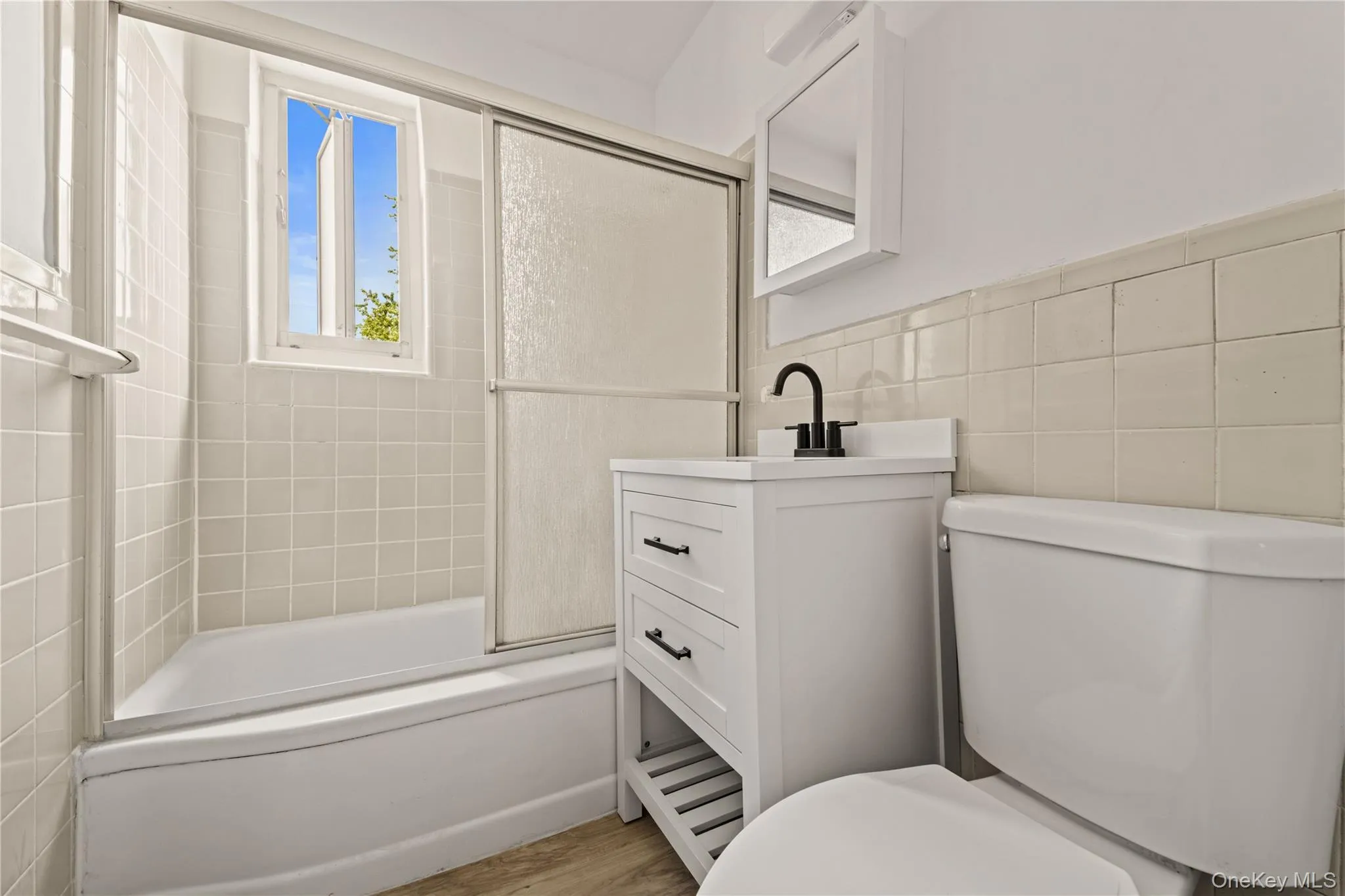 Full bath featuring tile walls, bath / shower combo with glass door, vanity, wainscoting, and wood finished floors Full bath featuring tile walls, bath / shower combo with glass door, vanity, wainscoting, and wood finished floors