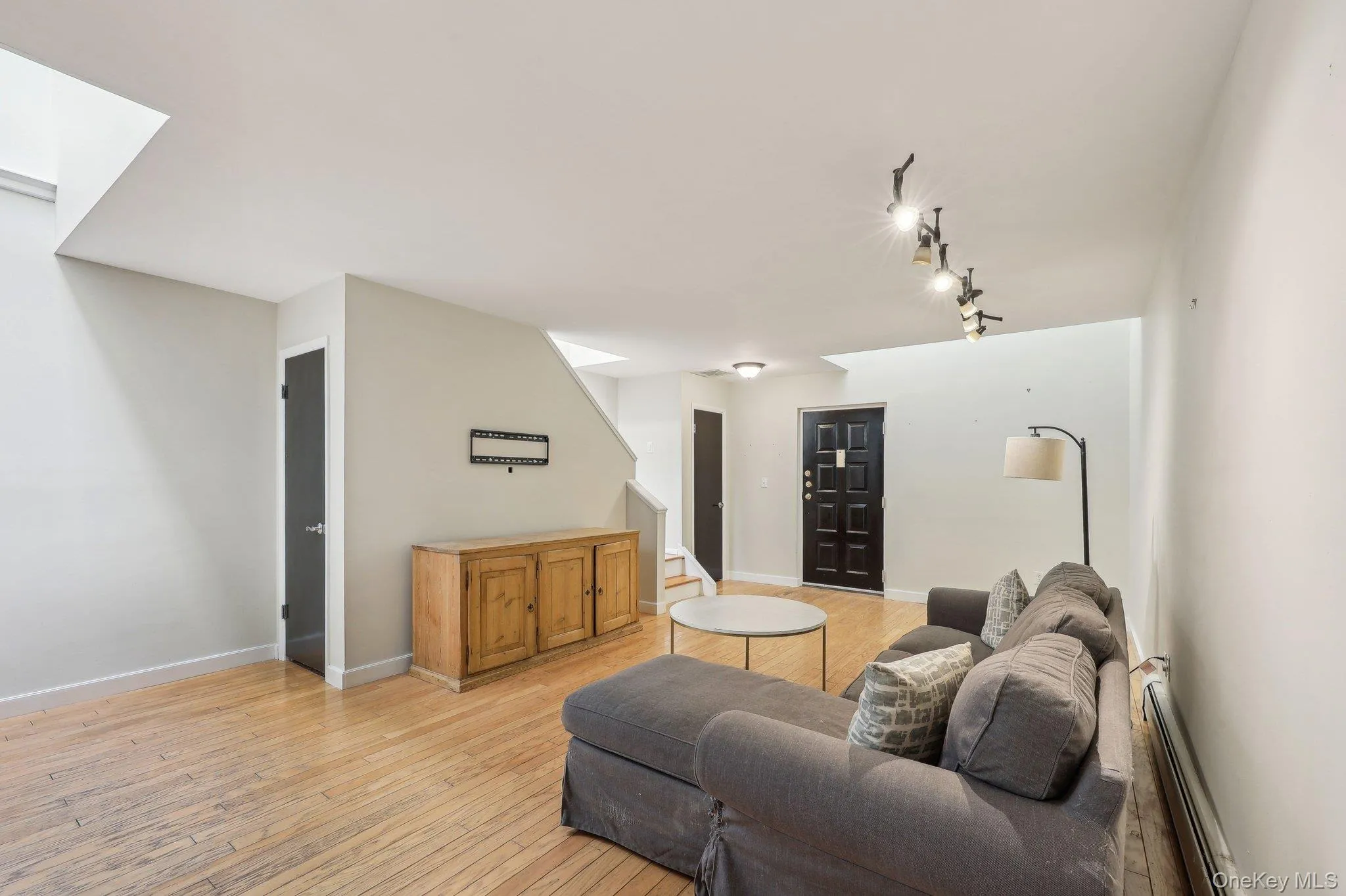 Living room with track lighting, light wood-style floors, baseboards, a baseboard radiator, and stairway Living room with track lighting, light wood-style floors, baseboards, a baseboard radiator, and stairway