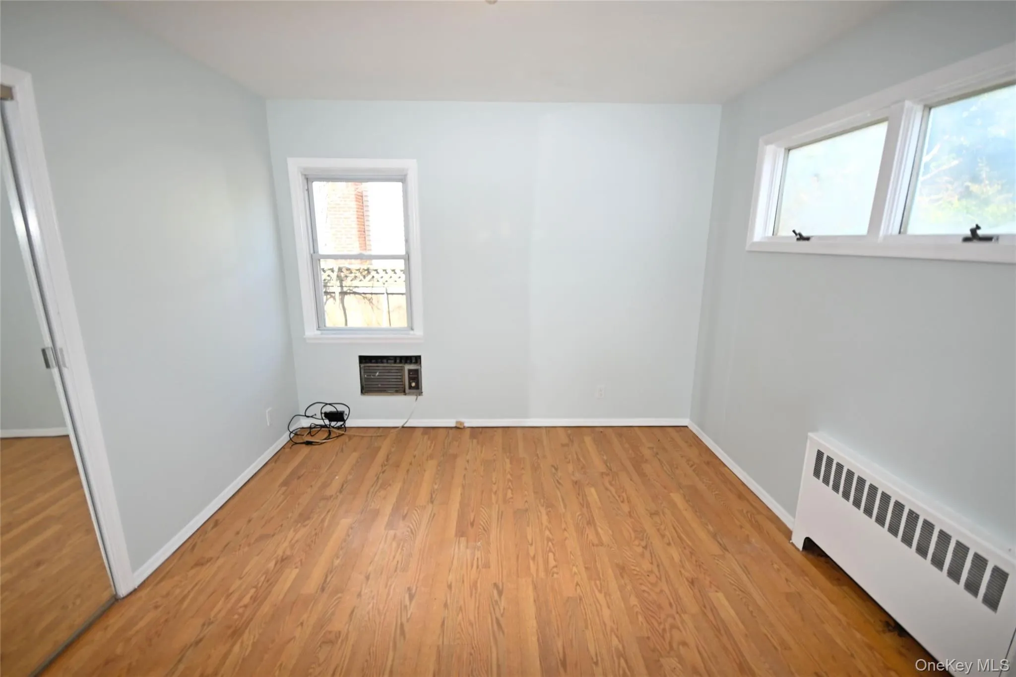 Unfurnished room featuring radiator heating unit, light wood finished floors, baseboards, and a wall unit AC Unfurnished room featuring radiator heating unit, light wood finished floors, baseboards, and a wall unit AC