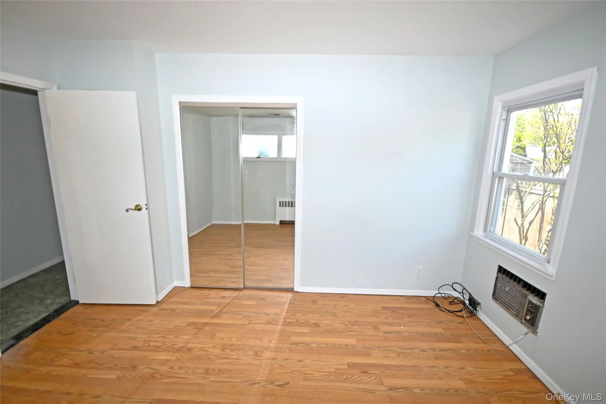 Unfurnished bedroom featuring radiator, light wood-type flooring, a wall mounted air conditioner, and baseboards Unfurnished bedroom featuring radiator, light wood-type flooring, a wall mounted air conditioner, and baseboards