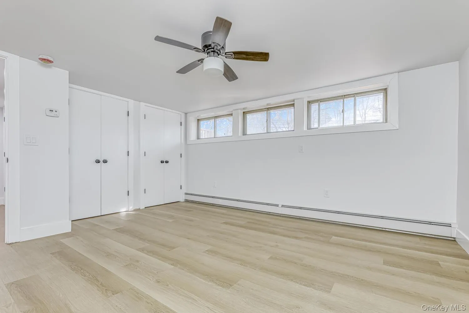 Unfurnished bedroom with two closets, baseboard heating, light wood-style flooring, baseboards, and ceiling fan Unfurnished bedroom with two closets, baseboard heating, light wood-style flooring, baseboards, and ceiling fan