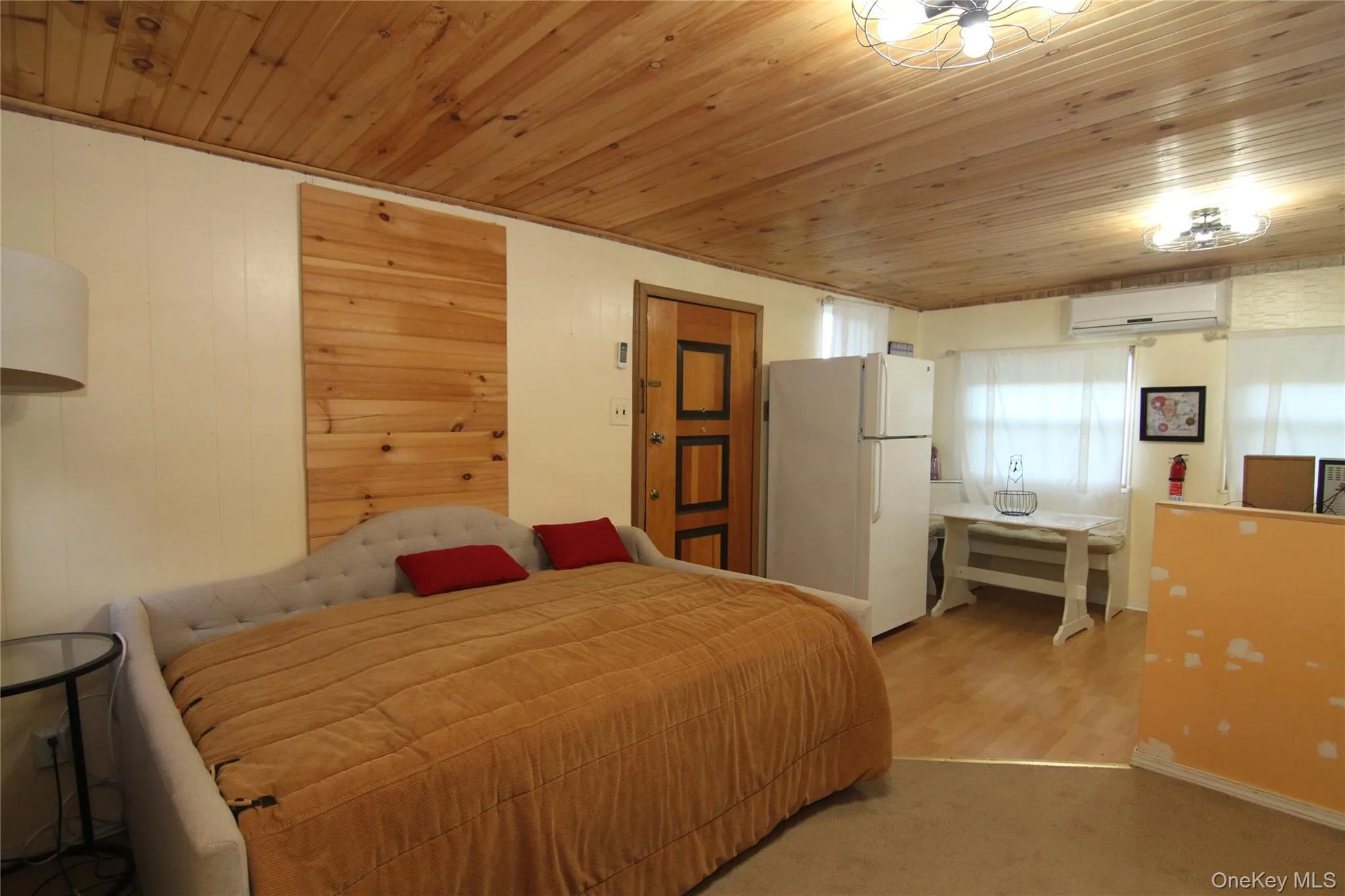 Carpeted bedroom with freestanding refrigerator, wooden ceiling, and a wall mounted air conditioner Carpeted bedroom with freestanding refrigerator, wooden ceiling, and a wall mounted air conditioner