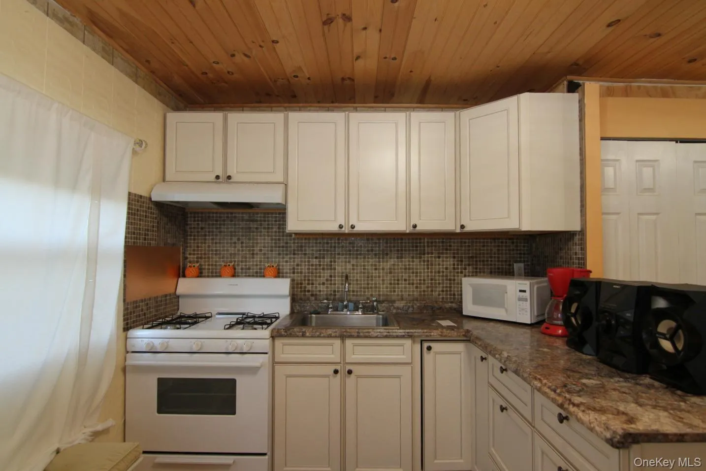 Kitchen with white appliances, a sink, under cabinet range hood, wooden ceiling, and backsplash Kitchen with white appliances, a sink, under cabinet range hood, wooden ceiling, and backsplash
