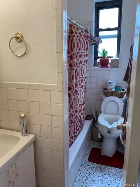 Full bath featuring tile walls, toilet, shower / bath combination with curtain, vanity, and wainscoting Full bath featuring tile walls, toilet, shower / bath combination with curtain, vanity, and wainscoting