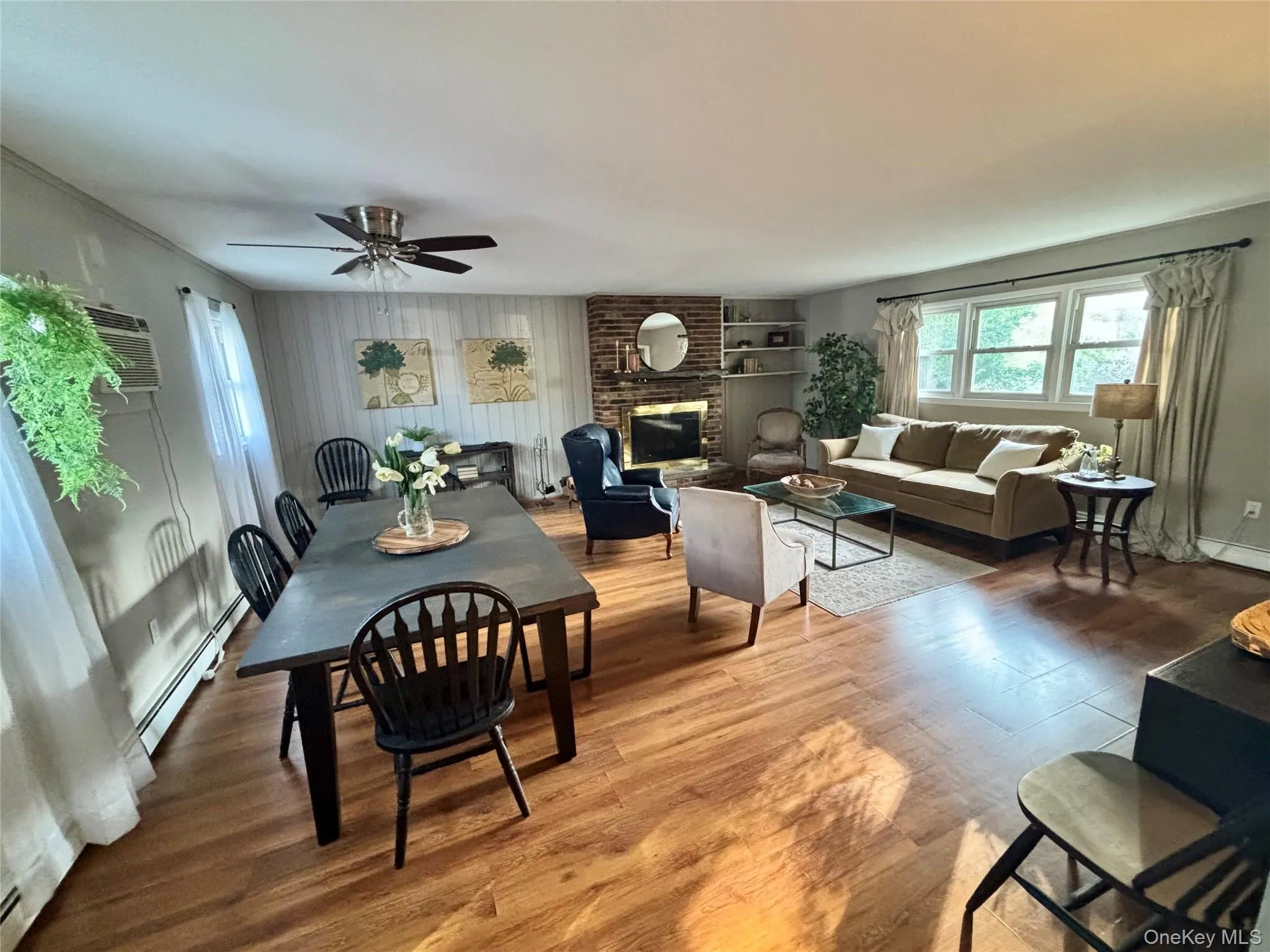Dining room featuring a baseboard heating unit, wood finished floors, ceiling fan, a wall mounted AC, and a brick fireplace Dining room featuring a baseboard heating unit, wood finished floors, ceiling fan, a wall mounted AC, and a brick fireplace