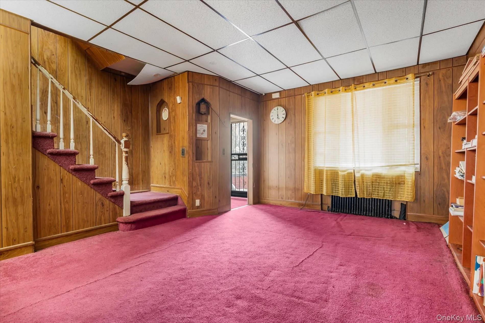 Carpeted living room featuring wood walls, stairway, baseboards, and a paneled ceiling Carpeted living room featuring wood walls, stairway, baseboards, and a paneled ceiling