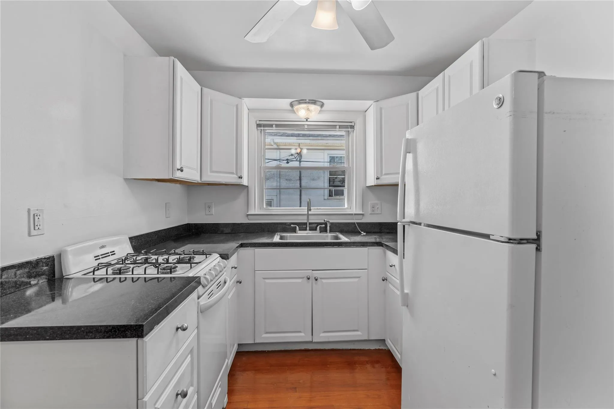 Kitchen with white appliances, a sink, dark countertops, a ceiling fan, and white cabinetry Kitchen with white appliances, a sink, dark countertops, a ceiling fan, and white cabinetry