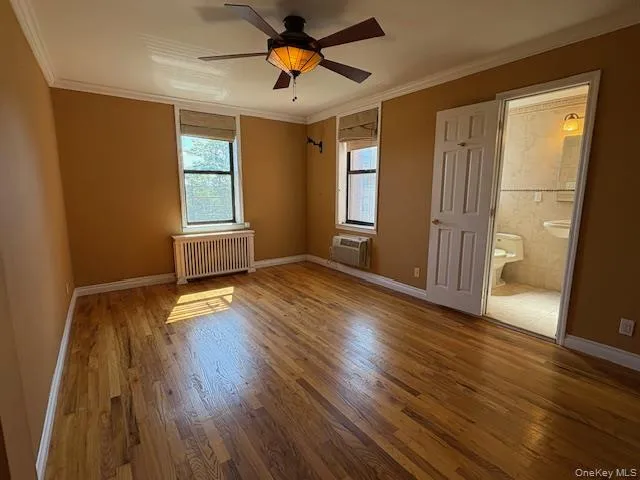 Unfurnished bedroom with crown molding, wood finished floors, multiple windows, radiator, and connected bathroom Unfurnished bedroom with crown molding, wood finished floors, multiple windows, radiator, and connected bathroom
