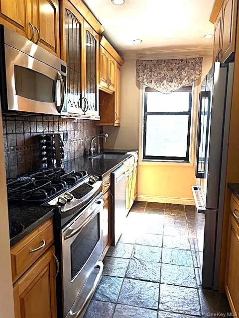 Kitchen featuring appliances with stainless steel finishes, glass insert cabinets, backsplash, brown cabinets, and stone tile floors Kitchen featuring appliances with stainless steel finishes, glass insert cabinets, backsplash, brown cabinets, and stone tile floors