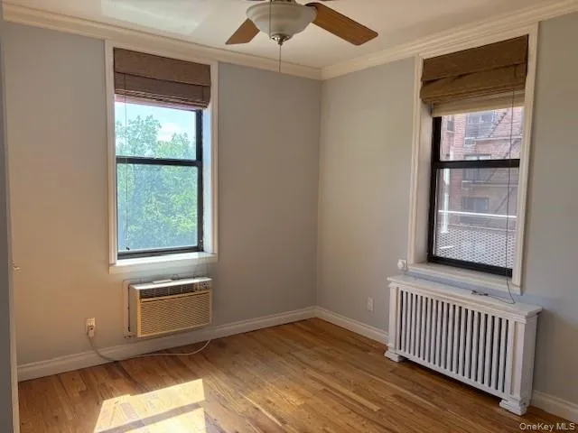 Spare room featuring radiator, light wood-style flooring, ornamental molding, ceiling fan, and a wall mounted air conditioner Spare room featuring radiator, light wood-style flooring, ornamental molding, ceiling fan, and a wall mounted air conditioner