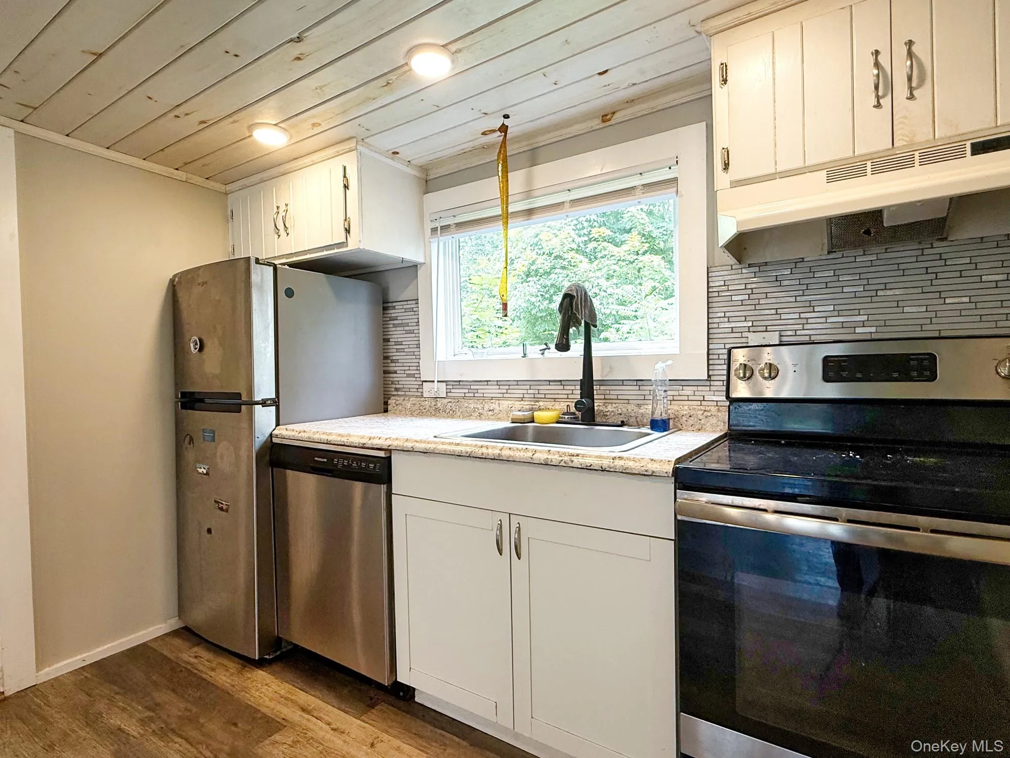 Kitchen featuring appliances with stainless steel finishes, under cabinet range hood, wood ceiling, wood finished floors, and backsplash Kitchen featuring appliances with stainless steel finishes, under cabinet range hood, wood ceiling, wood finished floors, and backsplash