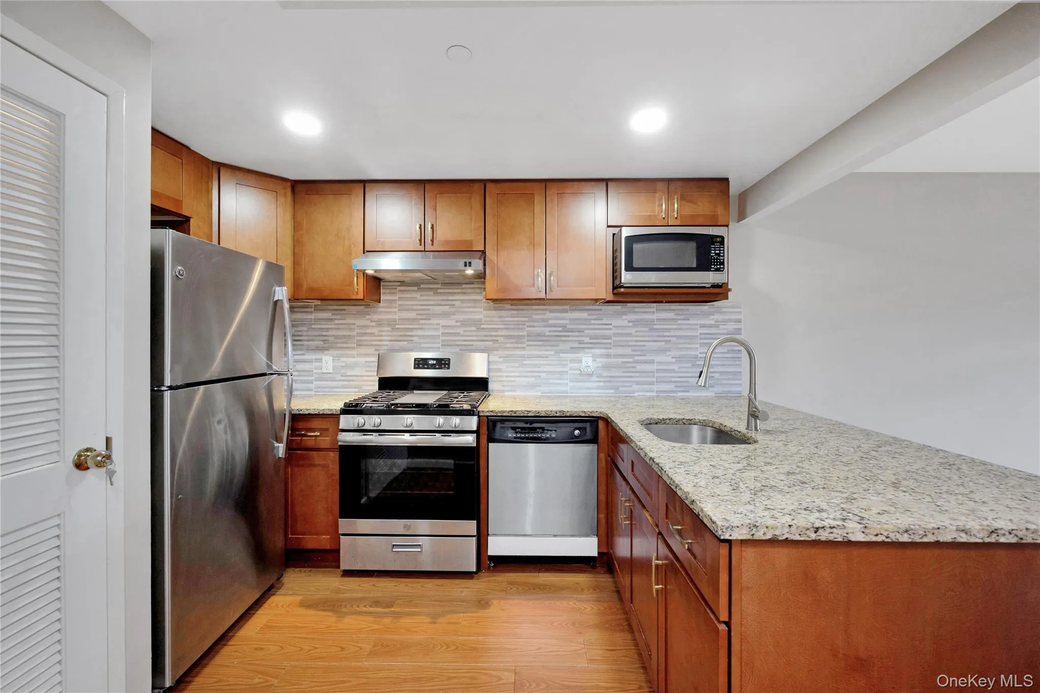 Kitchen featuring light wood-type flooring, under cabinet range hood, brown cabinetry, appliances with stainless steel finishes, and a sink Kitchen featuring light wood-type flooring, under cabinet range hood, brown cabinetry, appliances with stainless steel finishes, and a sink