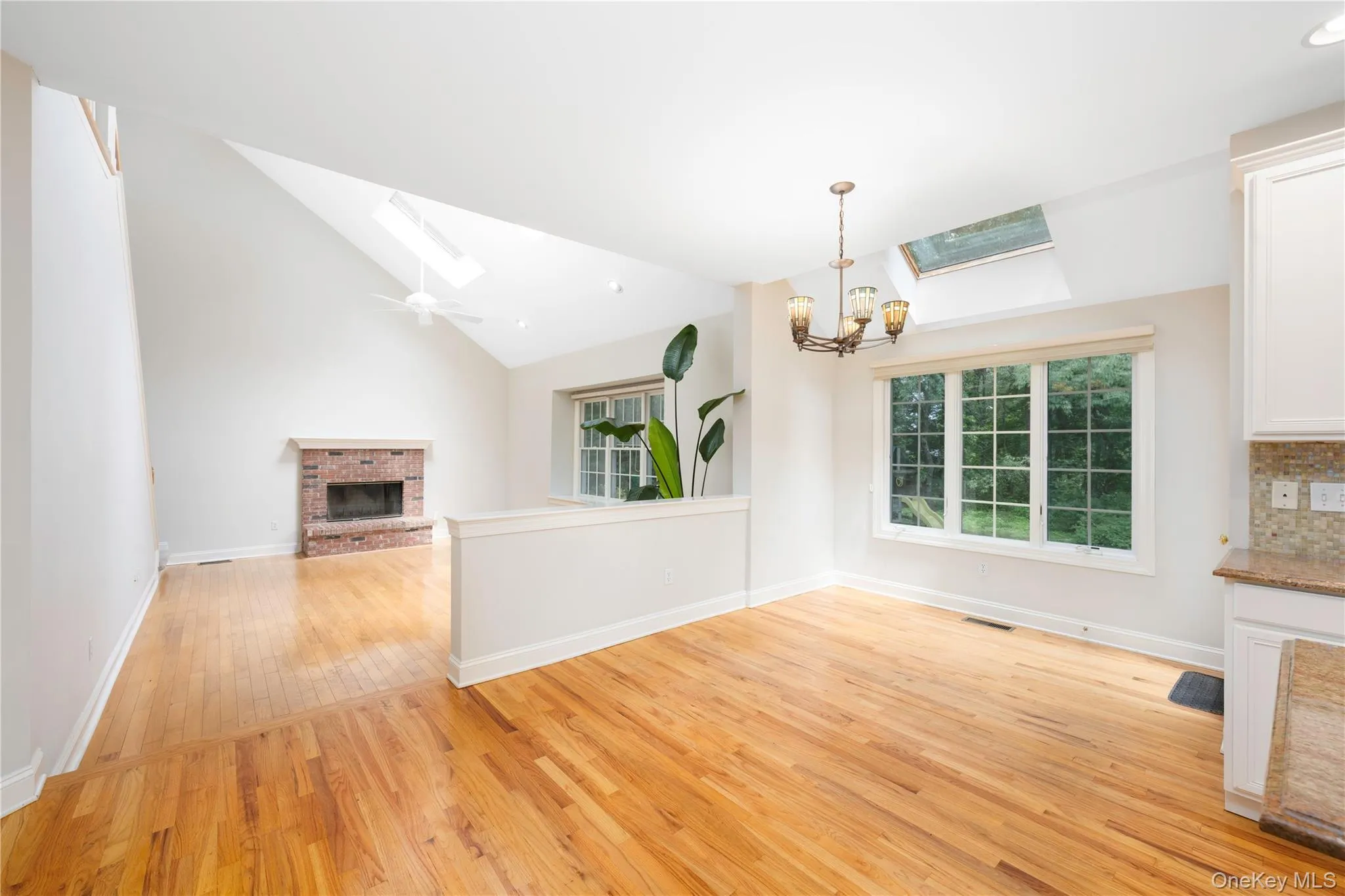 Unfurnished living room featuring a skylight, a brick fireplace, light wood-style flooring, a chandelier, and lofted ceiling Unfurnished living room featuring a skylight, a brick fireplace, light wood-style flooring, a chandelier, and lofted ceiling